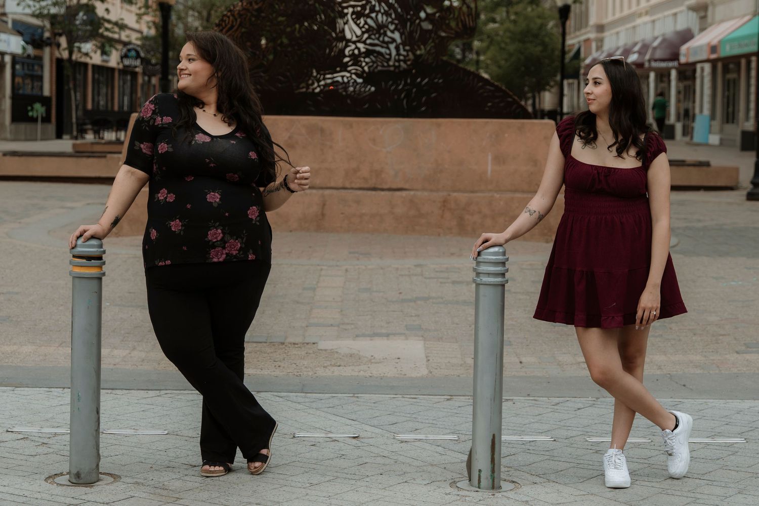 Two women posing outdoors near metal bollards on a brick plaza, one in black floral top and one in burgundy dress.