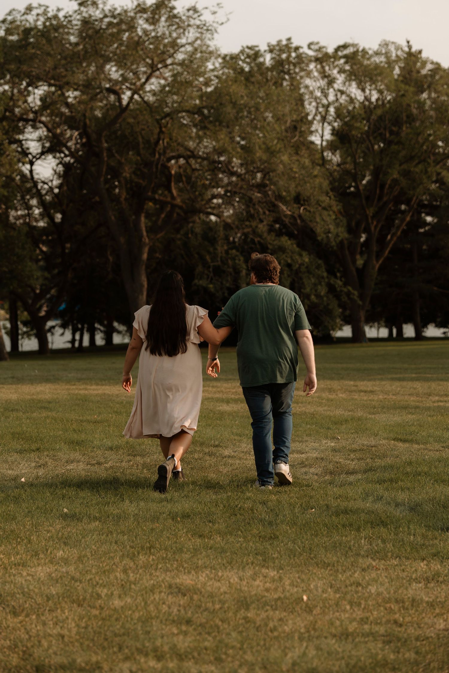 A couple walks hand-in-hand through a lush green park, surrounded by tall trees in warm evening light.
