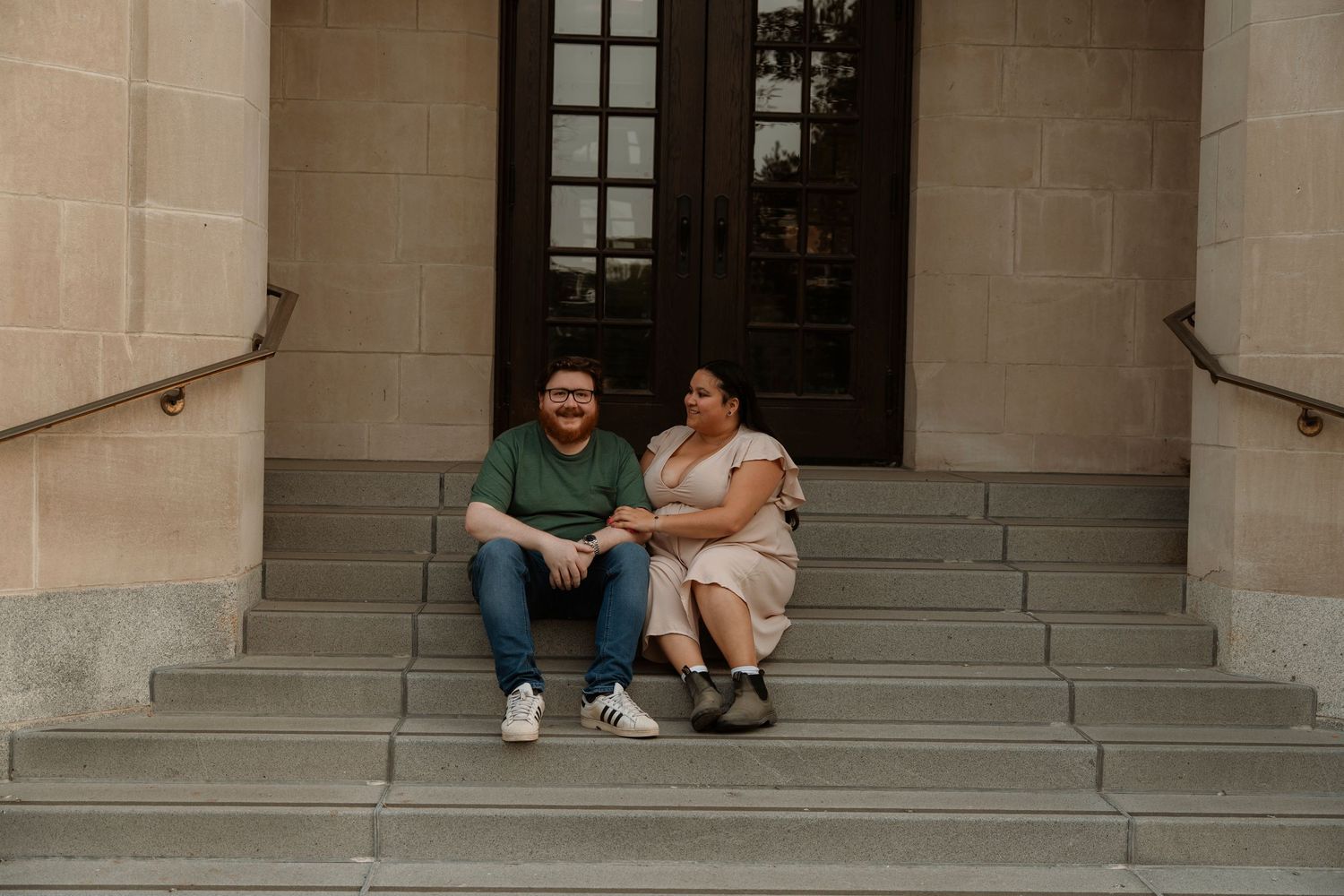 Couple sitting together on stone steps outside a historic building with large wooden doors, smiling and holding hands.
