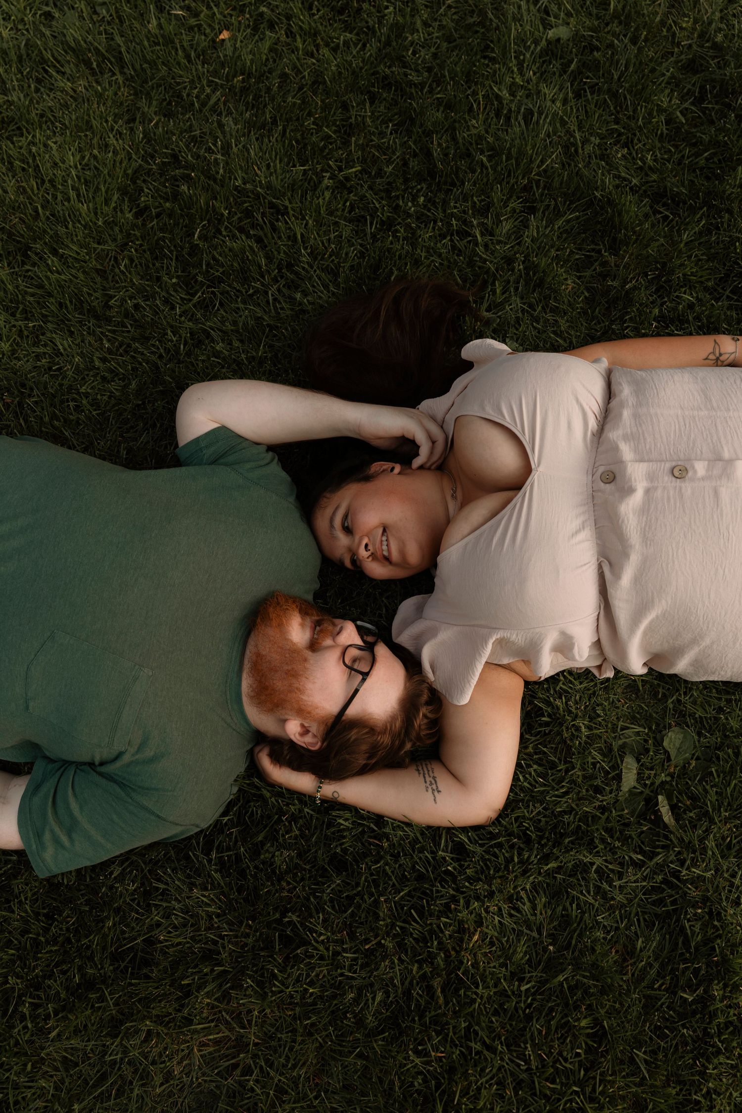 Couple lying on green grass looking at each other, man with red beard and glasses, woman in white dress.