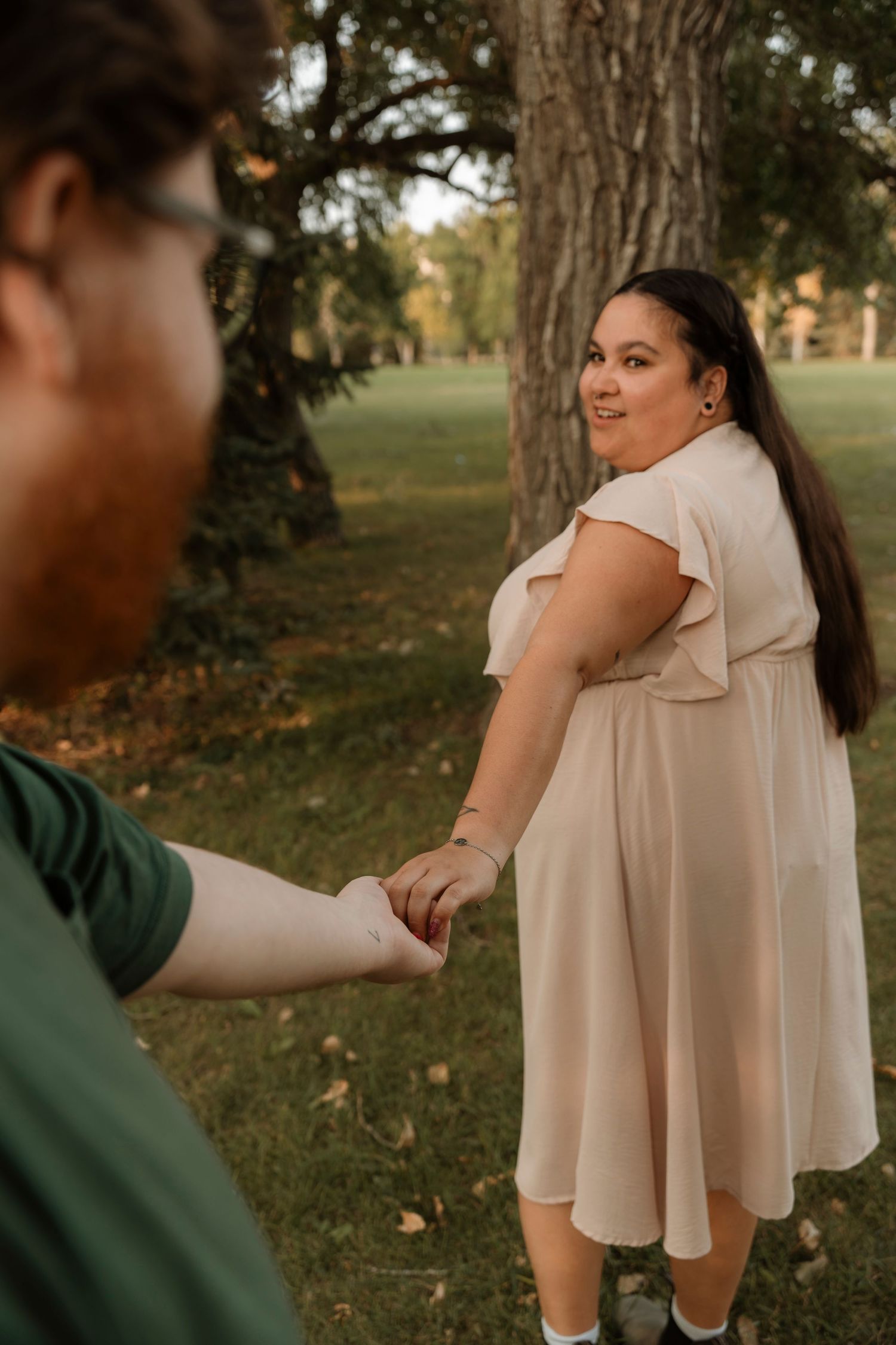 A woman in a blush midi dress holds hands with a man in a green shirt while looking back and smiling in a park.