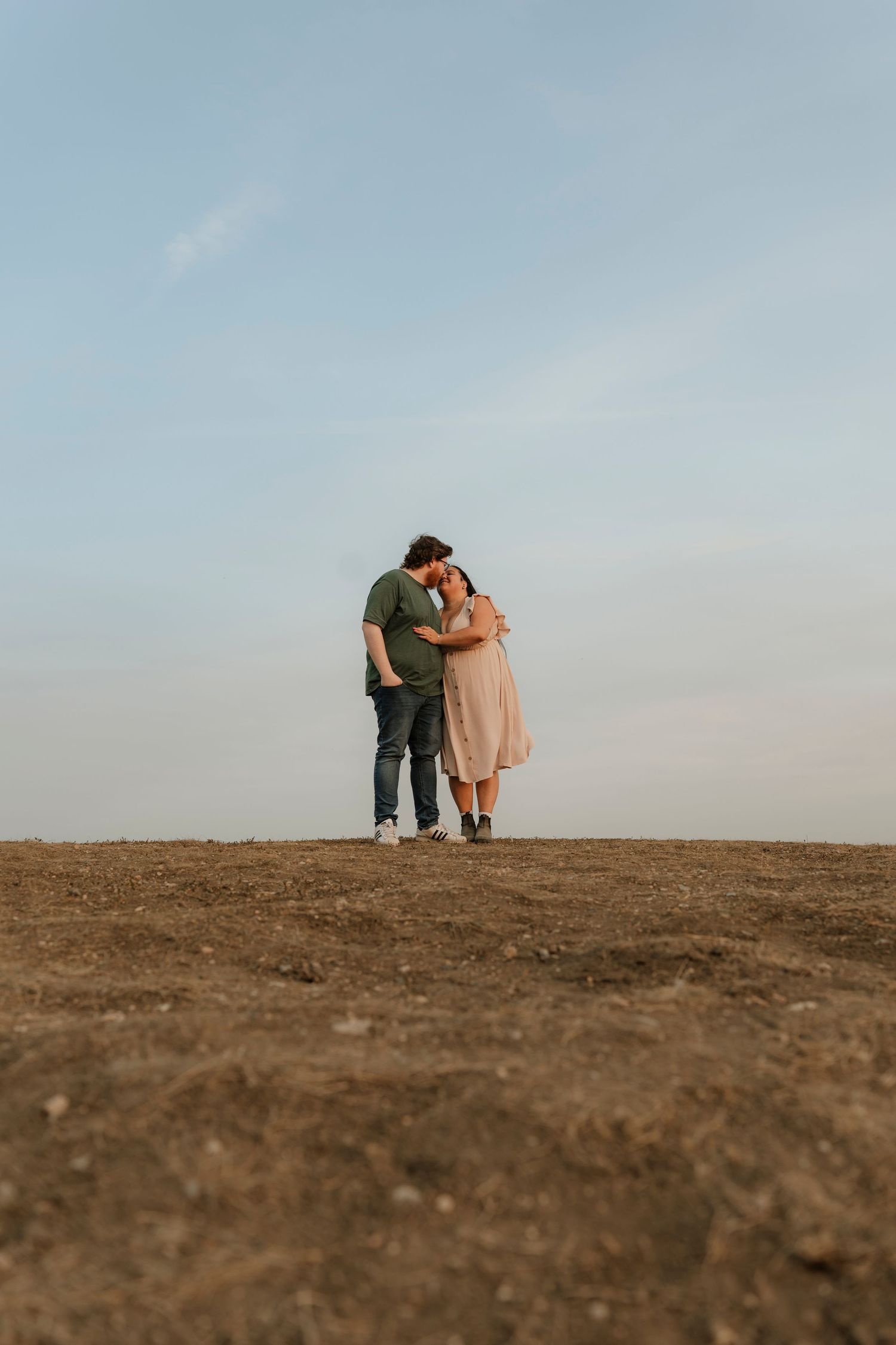A couple embraces tenderly on a barren hilltop under a soft blue sky in a romantic outdoor portrait.