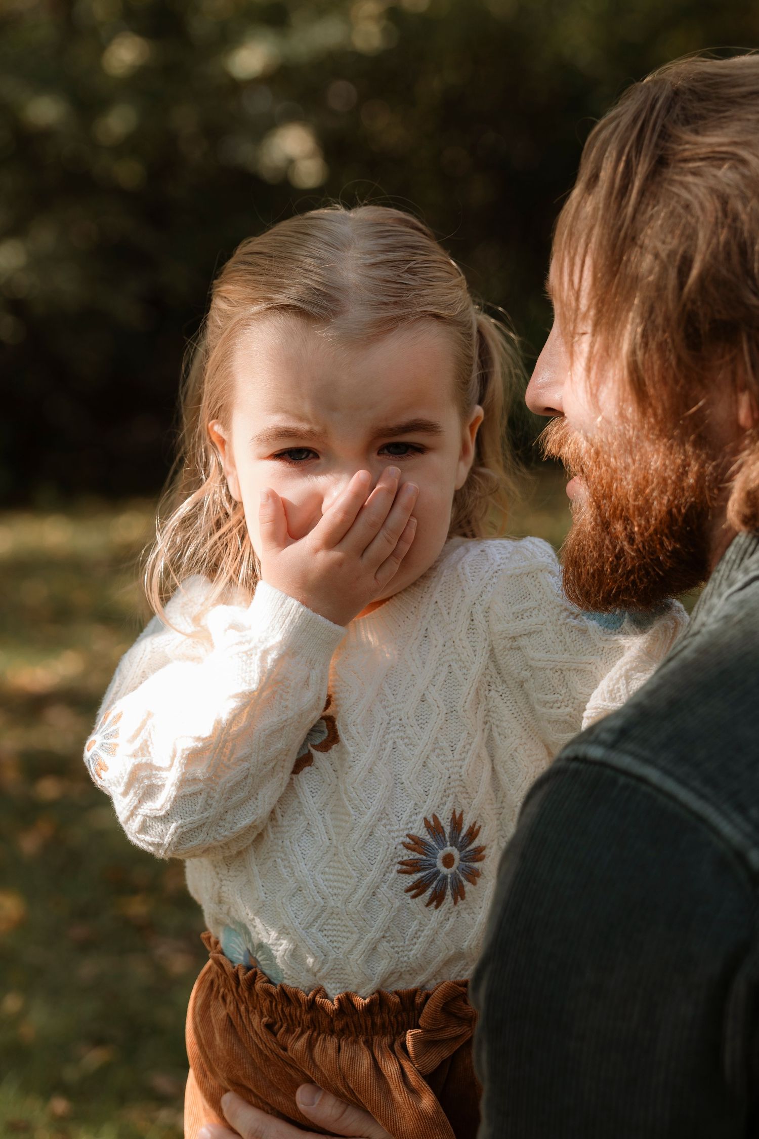 A bearded man holds a laughing young girl in a white knit sweater outdoors in a sunlit garden.