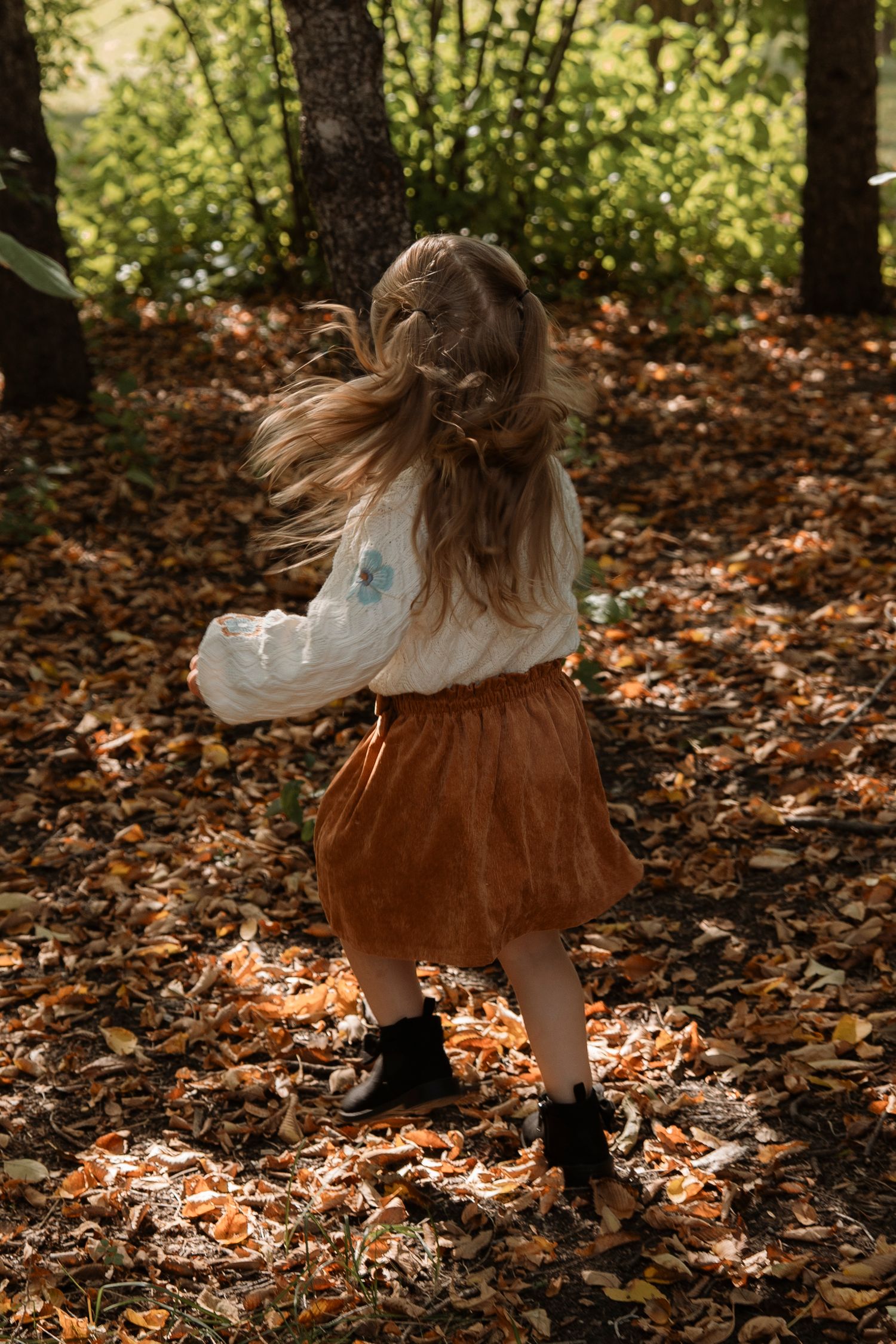 A young girl twirls in an autumn forest, wearing a white blouse and brown skirt, her long hair flowing.
