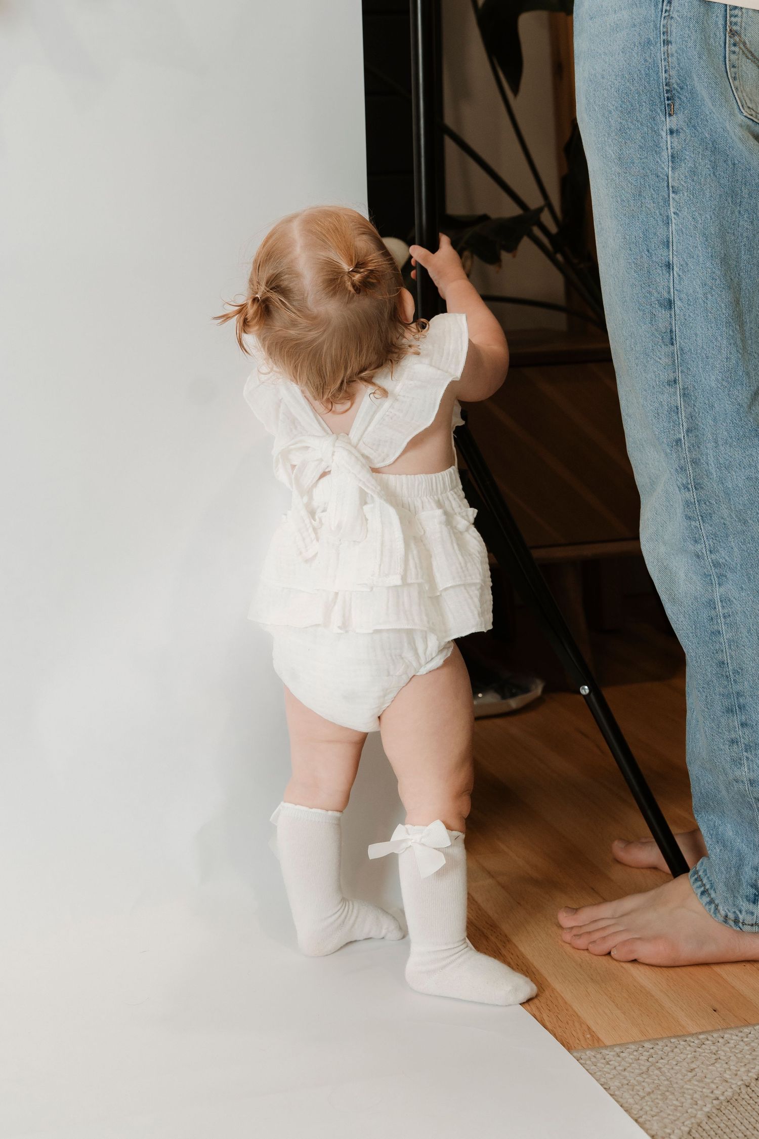 Toddler in white ruffled outfit and knee socks standing and holding a photography backdrop stand.