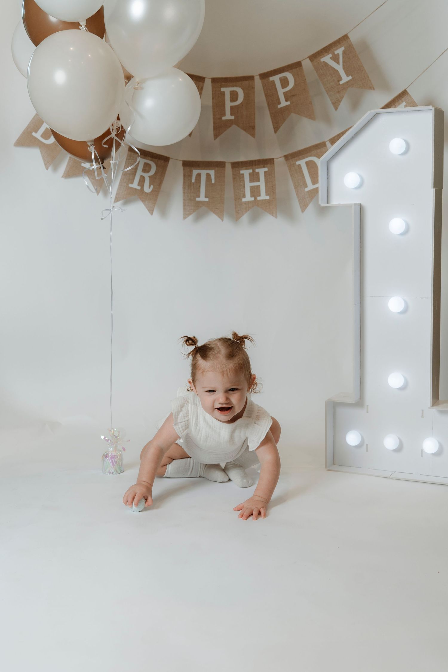 Smiling baby girl crawling on white floor at first birthday party with balloons and Happy Birthday banner.