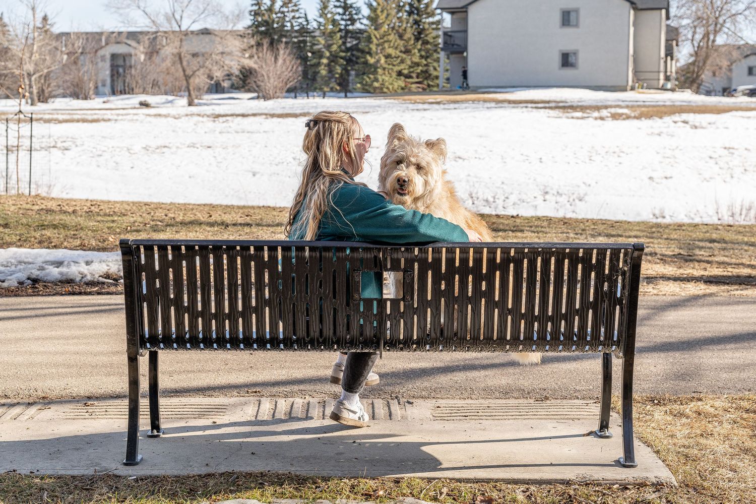 A woman and her fluffy dog sit together on a park bench in a snowy suburban setting.
