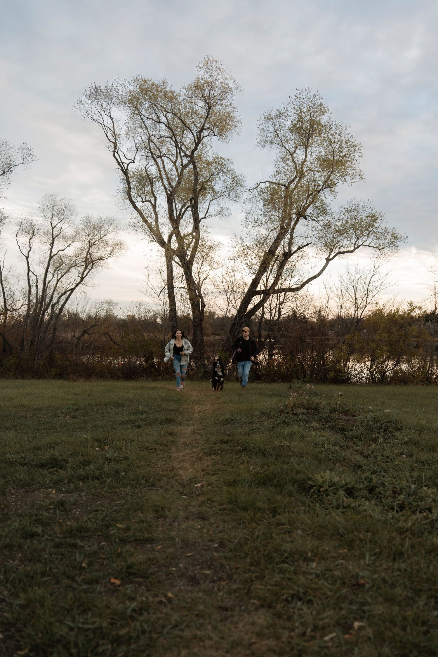 Two women walking a dog on a grassy path in an autumn park with bare trees and cloudy sky.