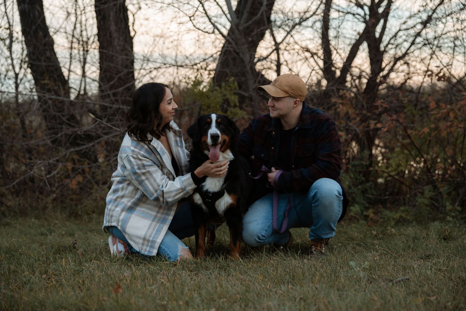 Couple crouching outdoors with a Bernese Mountain Dog in a wooded autumn setting.