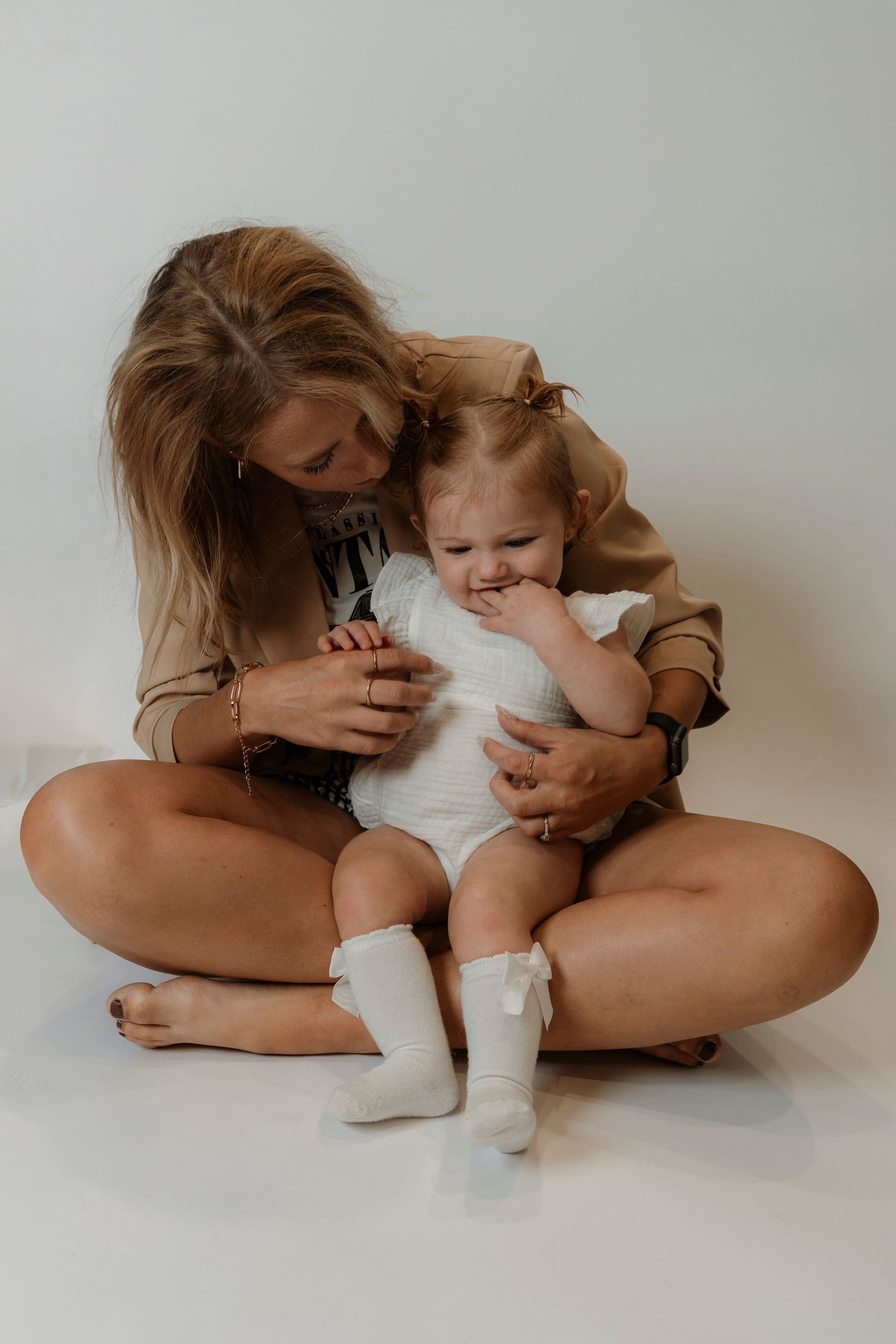A young woman sits cross-legged on a white floor, lovingly cradling a baby in a white onesie and socks.