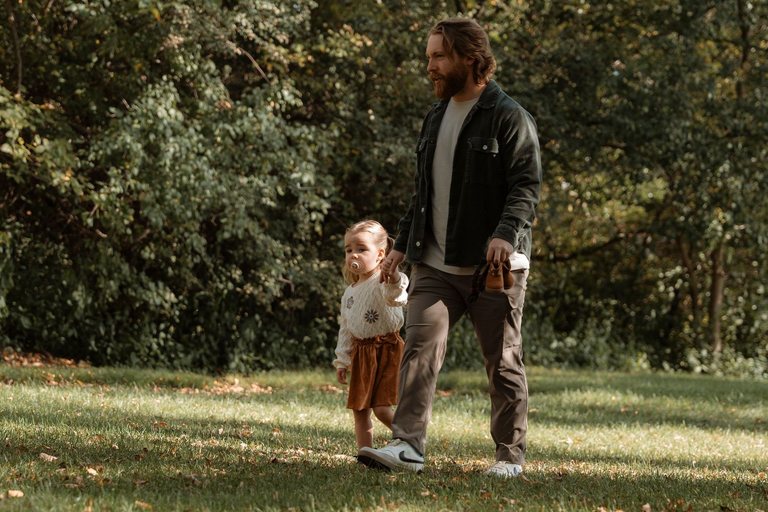 A bearded father walks with his toddler on green grass in a sunlit park surrounded by lush trees.