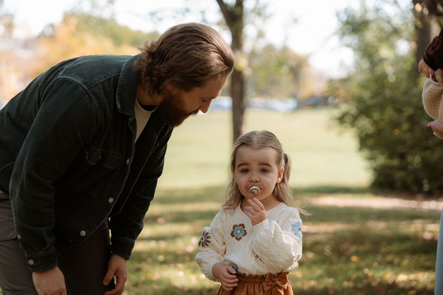 A father bends down toward his young daughter blowing a dandelion in a sunny park setting.