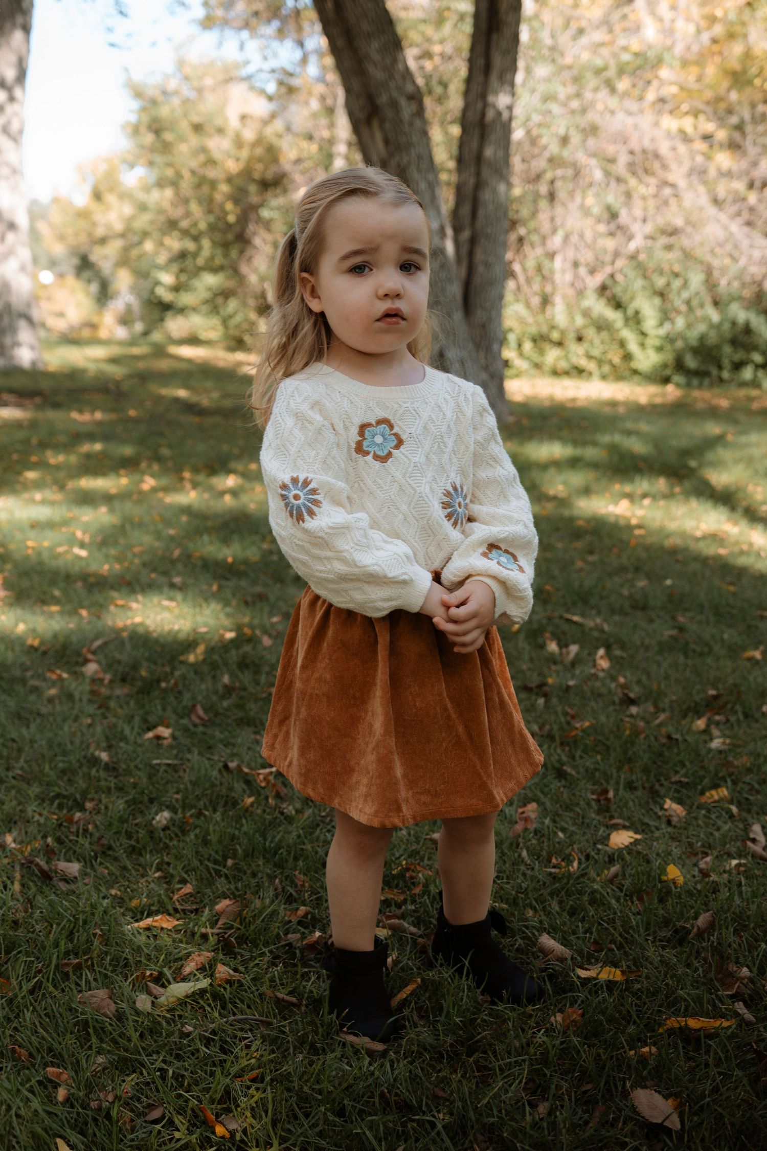 Toddler girl in white floral sweater and rust corduroy skirt with black boots standing on green grass outdoors.