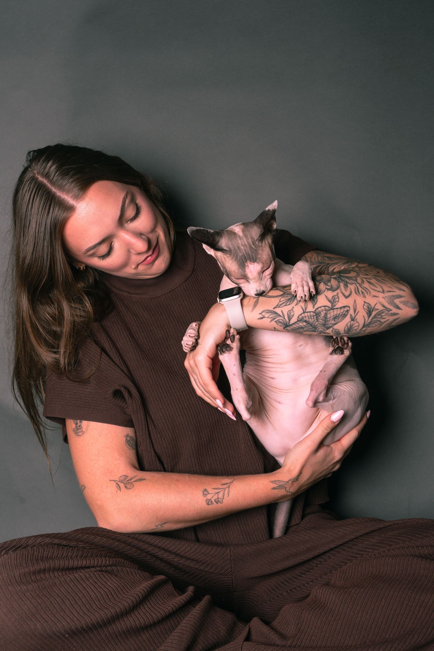 Tattooed woman in brown outfit holds a hairless Sphynx cat against a dark gray background.