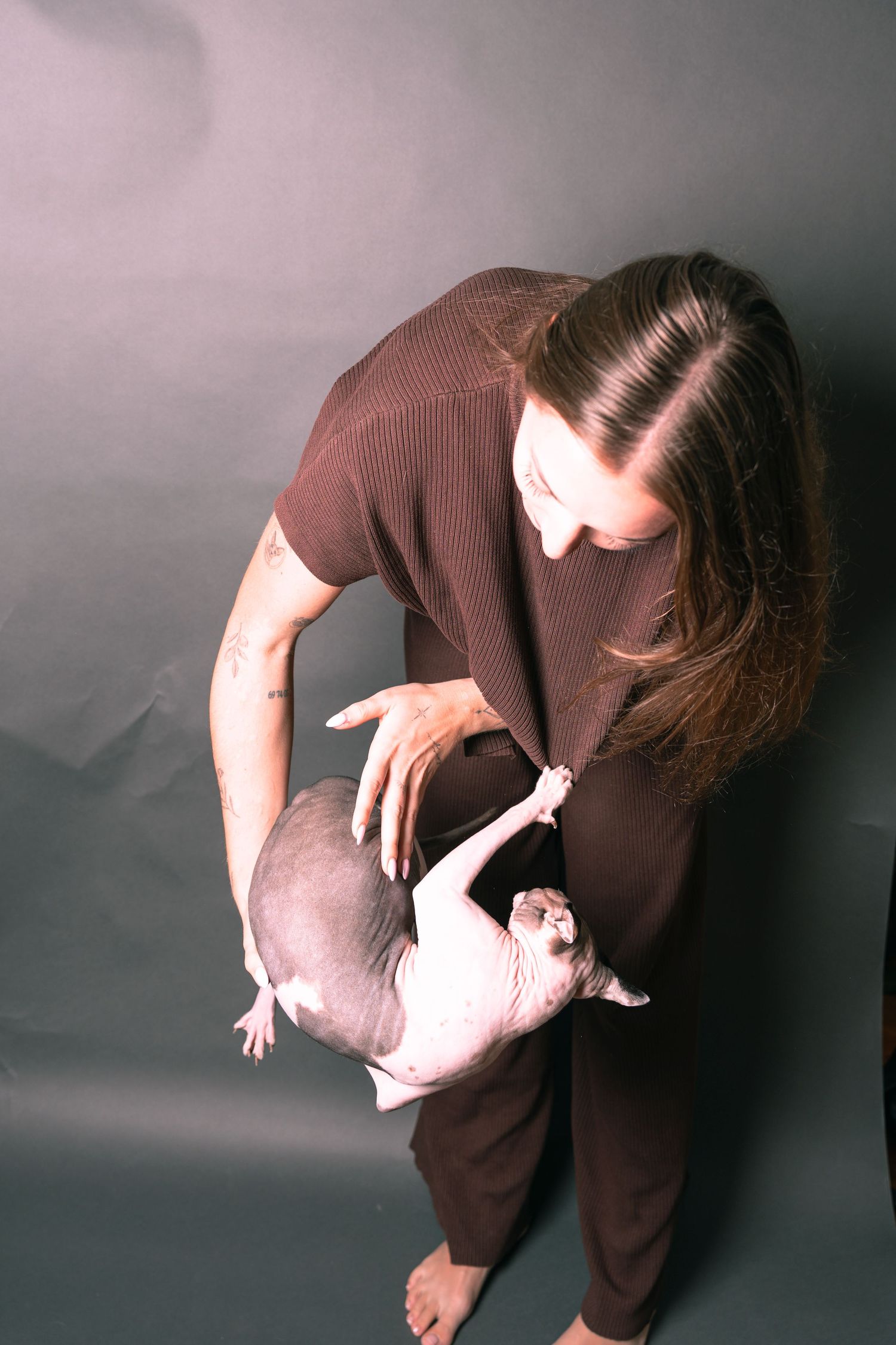 Woman in brown outfit holding a hairless Sphynx cat against a dark gray studio background.
