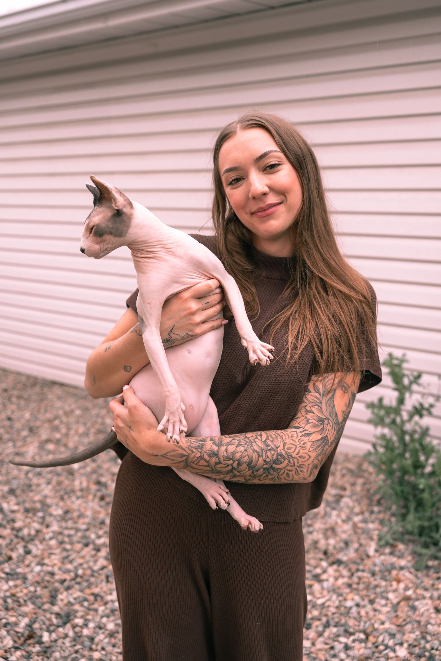 Tattooed woman in brown outfit smiling while holding a hairless Sphynx cat outdoors near a white building.