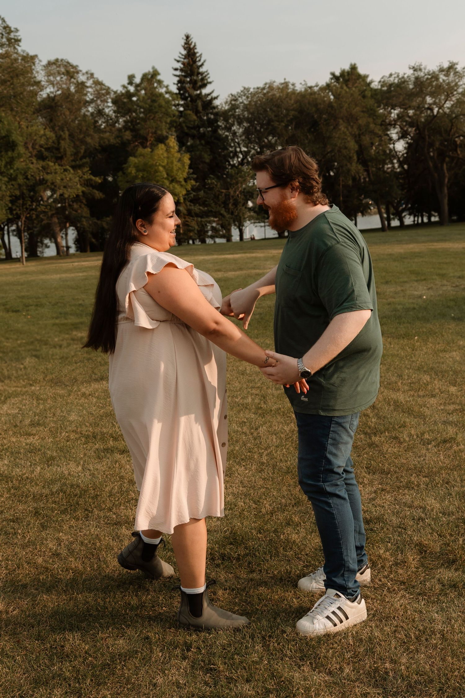 Couple holding hands and smiling at each other in a sunny park, woman in pink dress, man in green shirt and jeans.