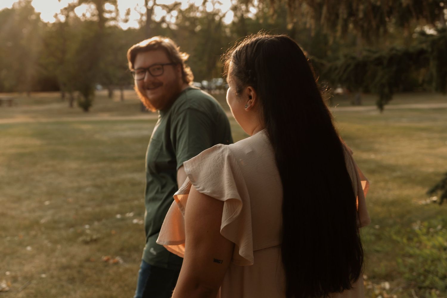 Couple sharing a golden hour moment outdoors, woman in ruffled dress and man in green shirt smiling in a park.