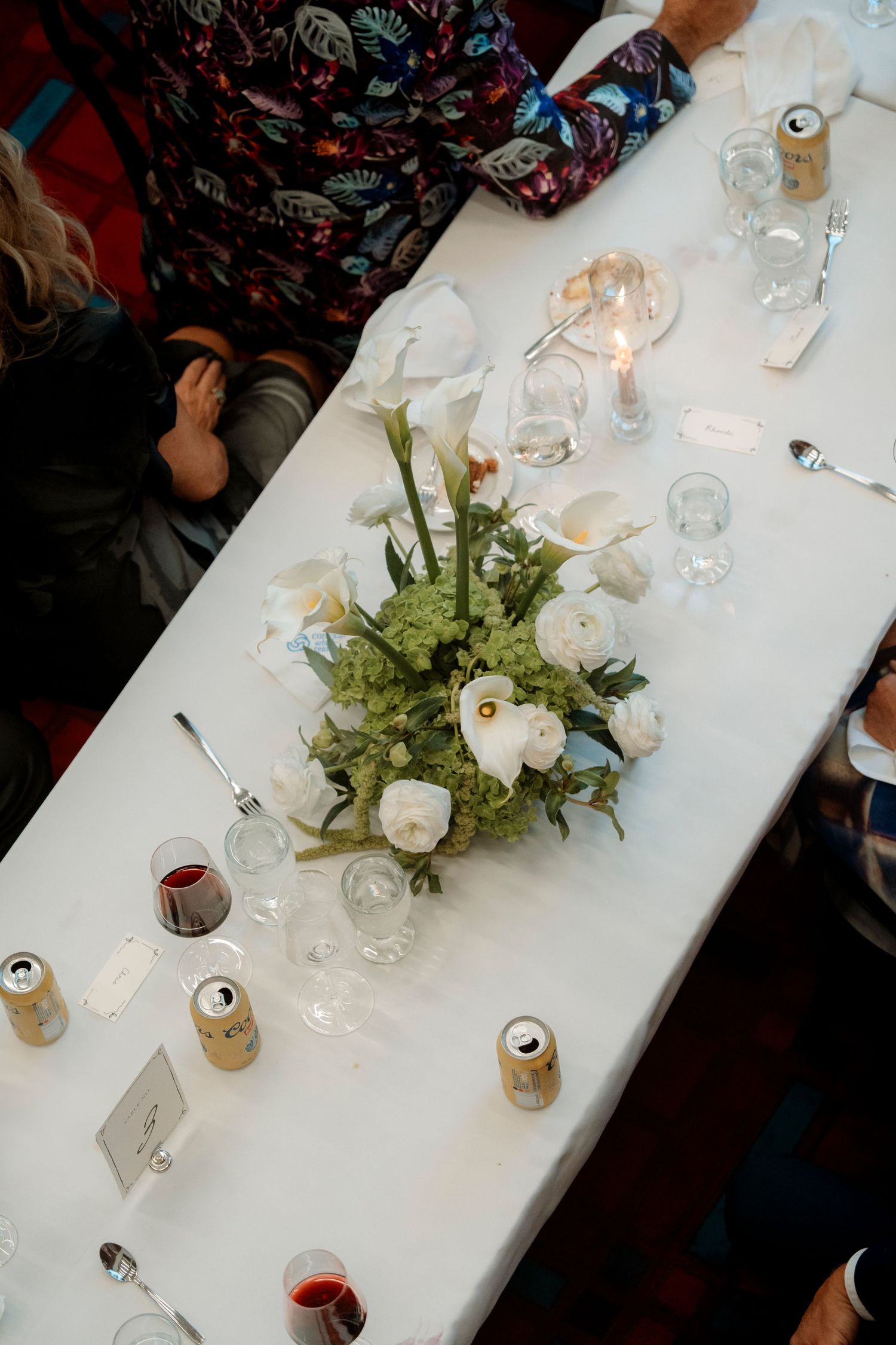 Elegant wedding reception table with white floral centerpiece, candles, wine glasses, and canned beverages on white linen.