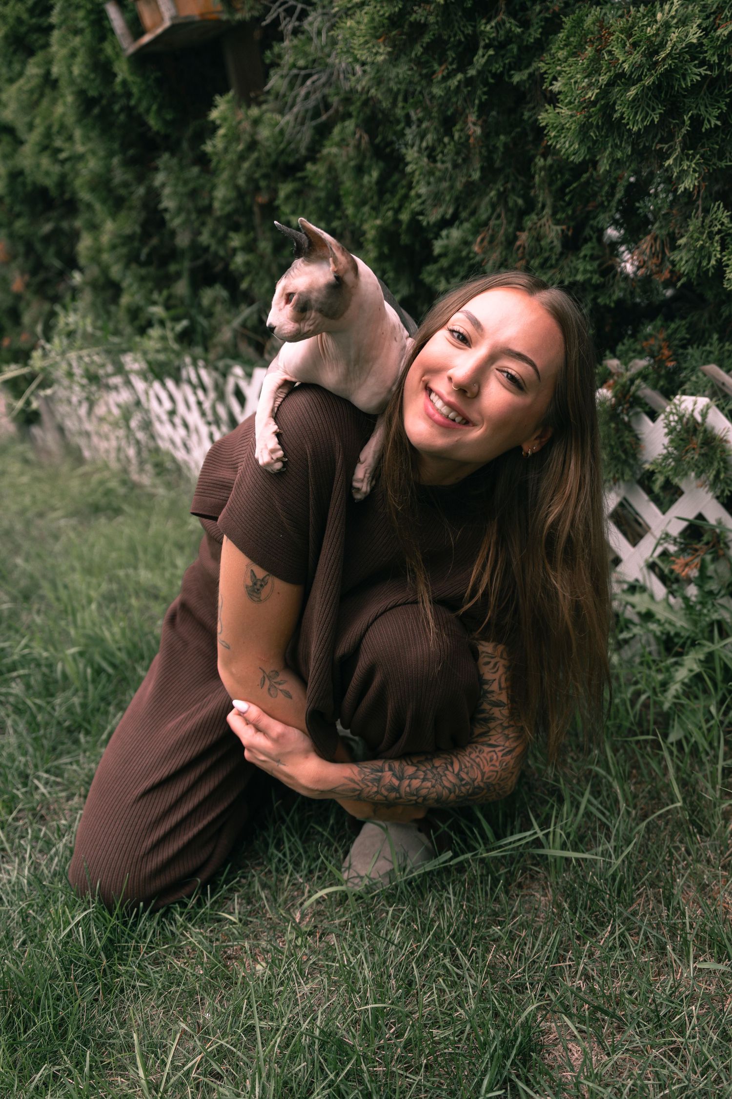 Smiling woman with tattoos crouching on grass, holding a small cat on her shoulder near green bushes.