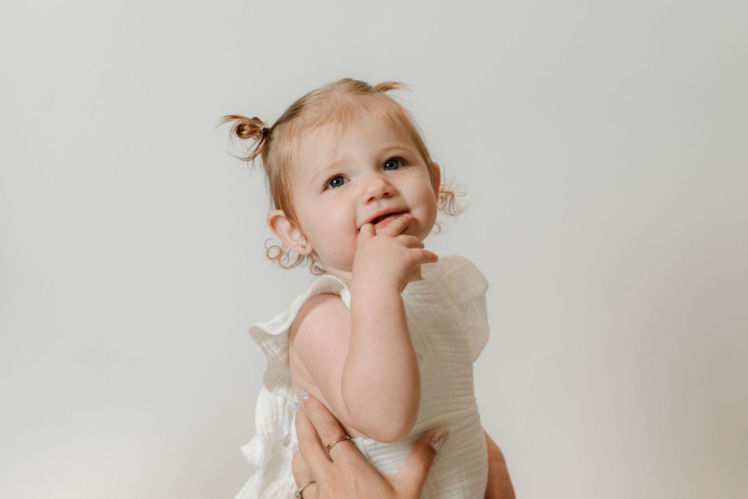 Adorable baby girl in white dress with tiny pigtails, smiling and touching her face against a light background.