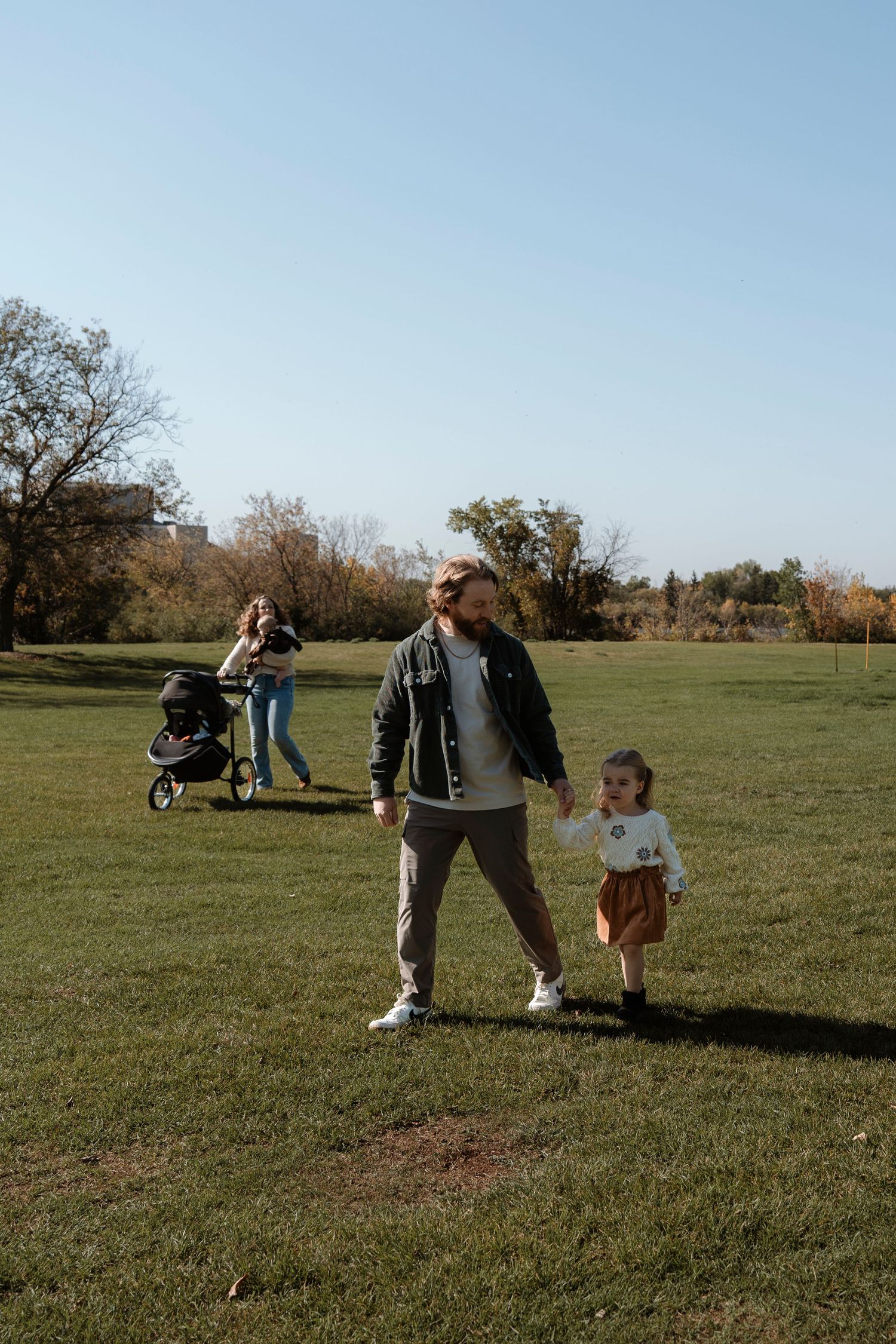 A family enjoys a sunny day in a green park, with a man holding a toddler's hand and a woman pushing a stroller.