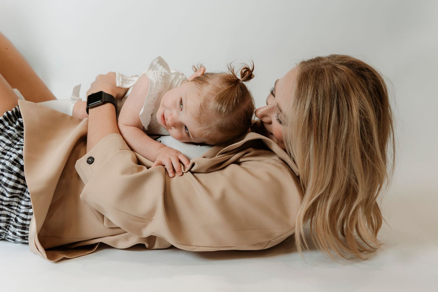 A blonde mother lying down tenderly gazing at her smiling baby in a white dress on a light background.