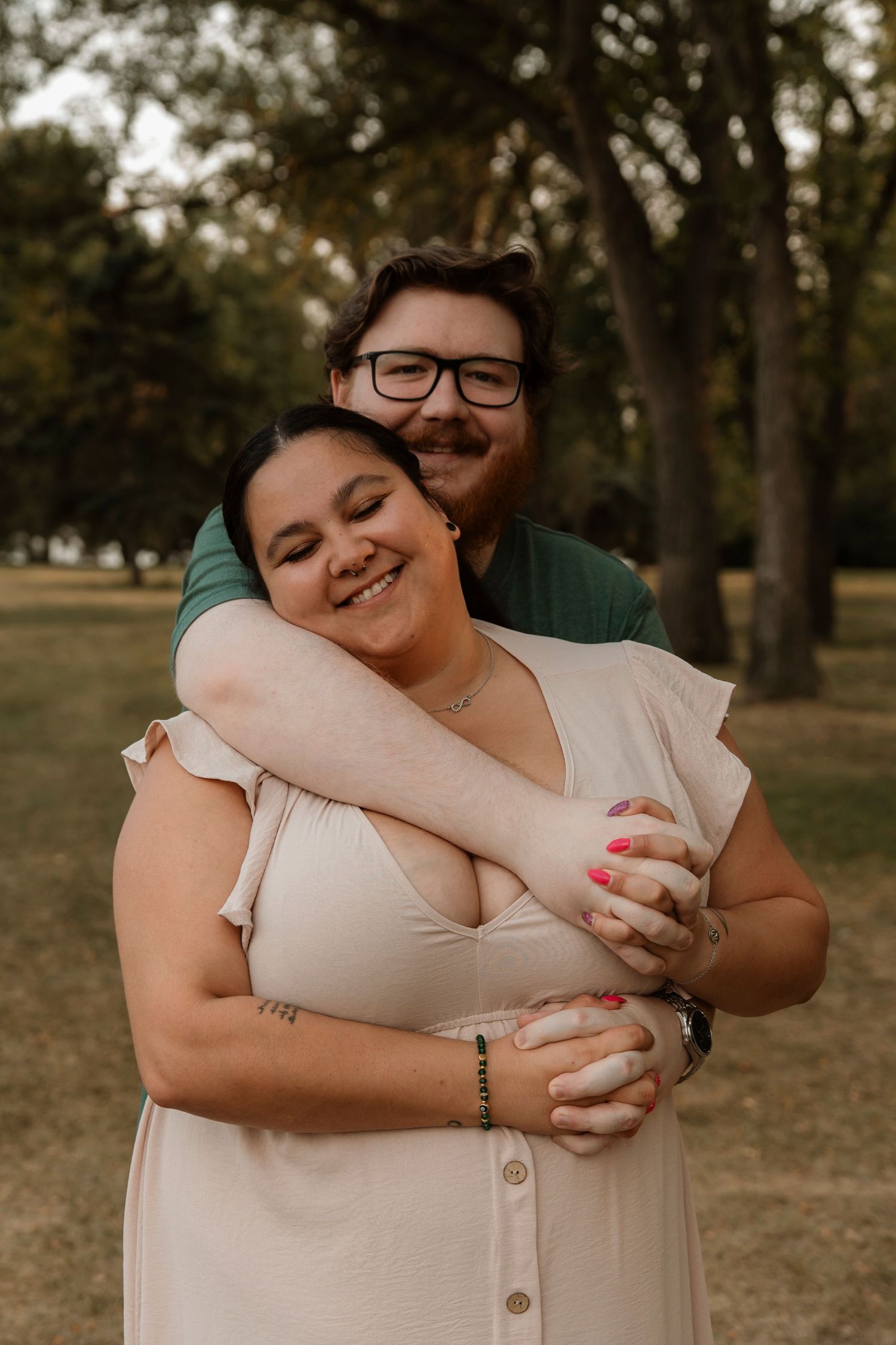 A couple embraces warmly in a park, the woman smiling joyfully in a white dress while the man hugs her from behind.