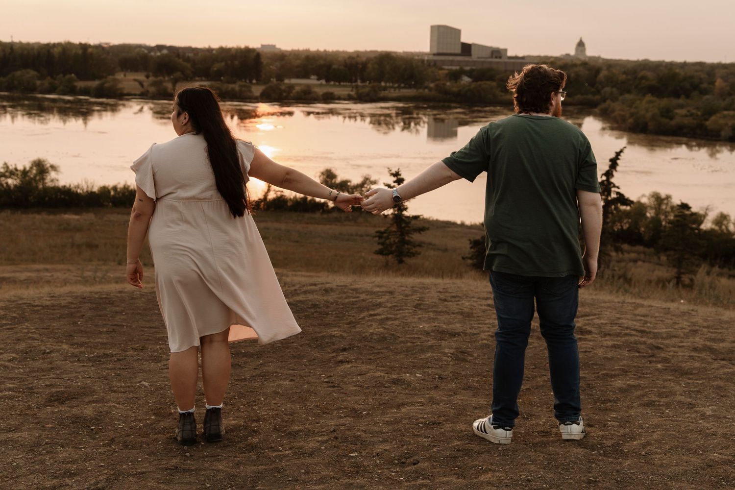 Couple holding hands at sunset overlooking a river, woman in white dress and man in green shirt, romantic outdoor scene.