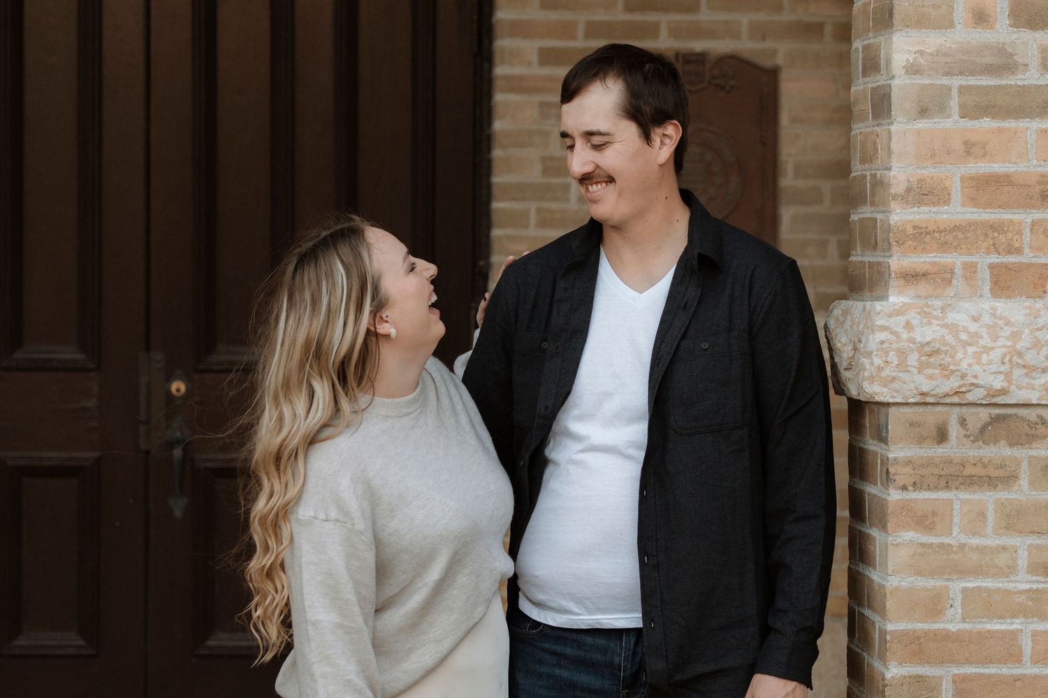 A couple smiles at each other standing by a brick wall and wooden door in a warm, candid outdoor portrait.