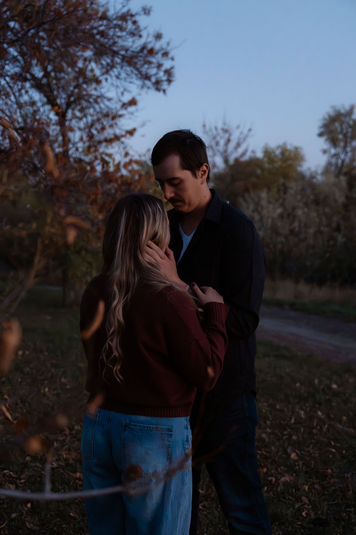 A couple embraces tenderly at dusk in an autumn park, the woman in a burgundy sweater and jeans, the man in dark clothing.