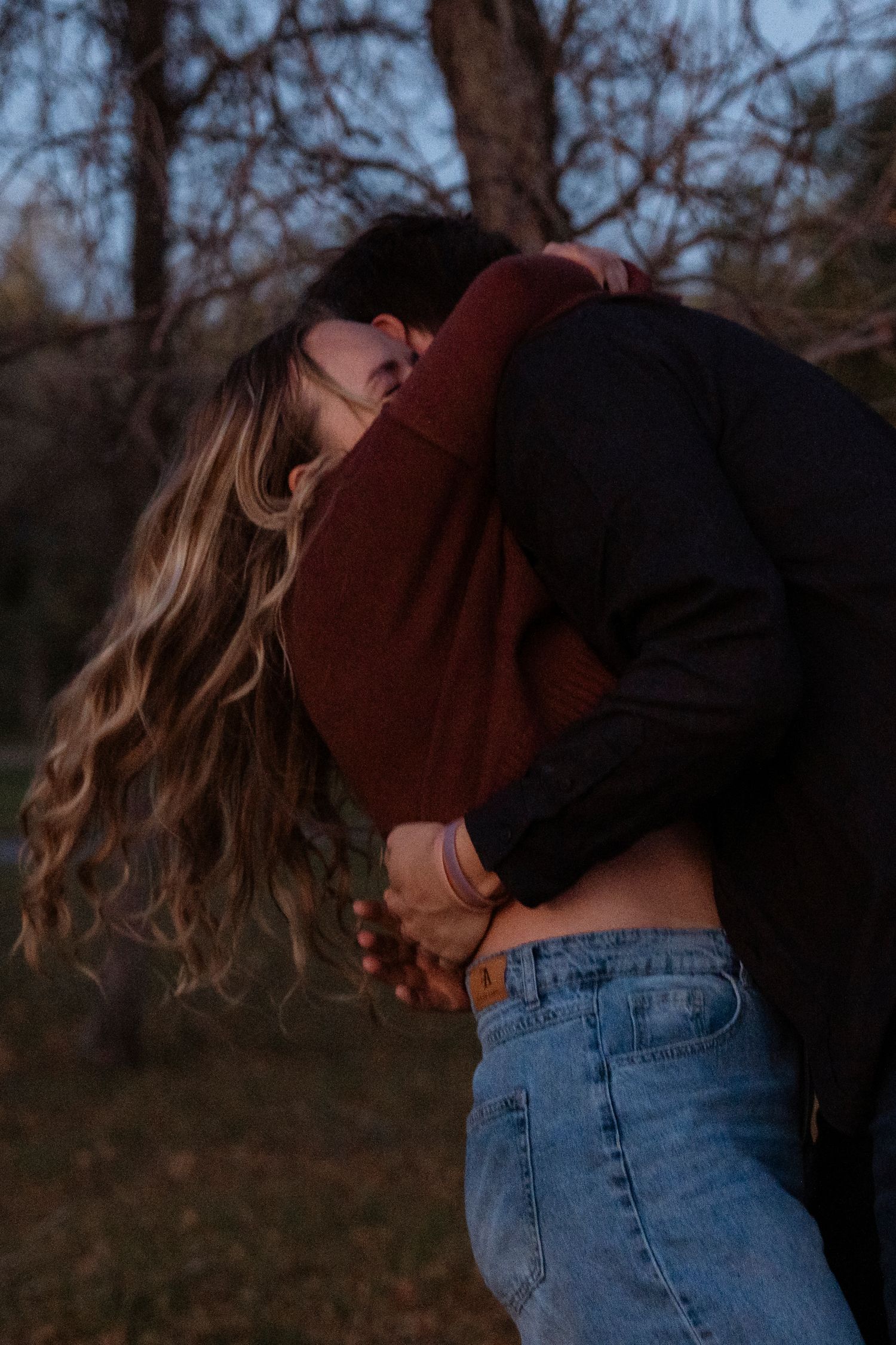 Couple embracing and kissing outdoors near bare trees at dusk, woman with long wavy hair in blue jeans.