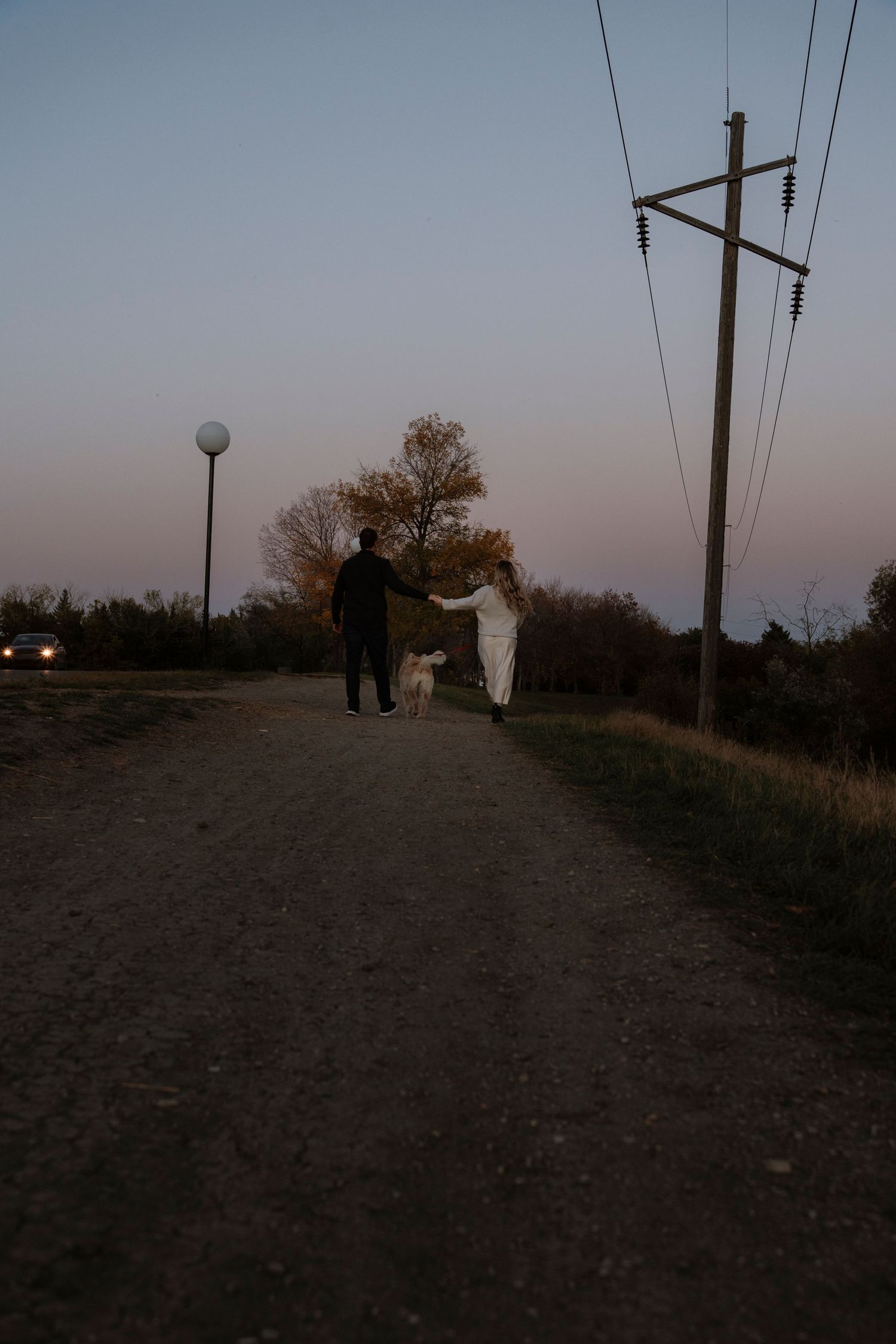 Couple walking a white dog down a rural dirt road at dusk, with power lines and a street lamp nearby.