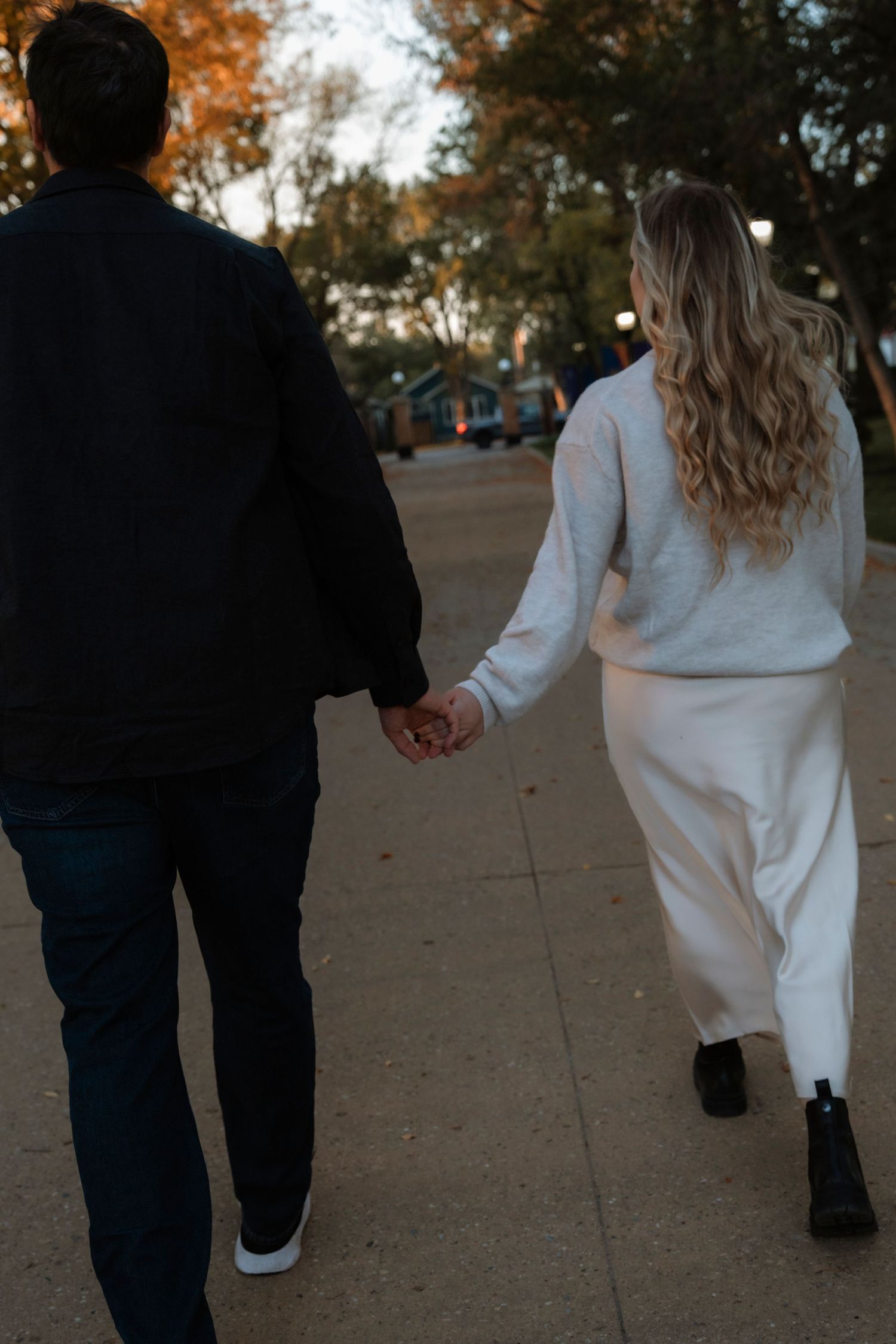 A couple holds hands while walking along a sidewalk in autumn, woman in white maxi skirt and gray sweater.