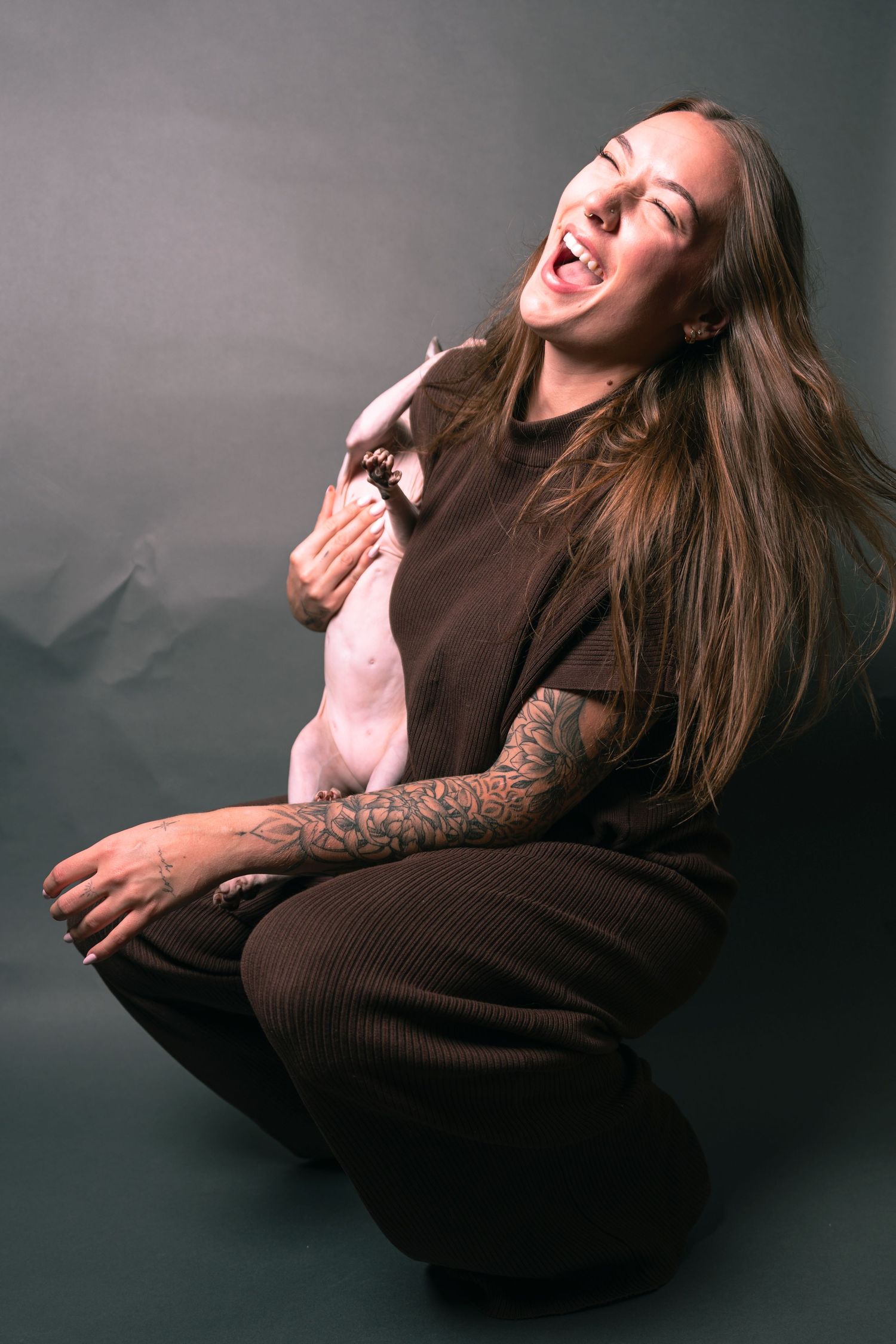A laughing woman with long hair and tattoos sits on the floor holding a Sphynx cat against her brown outfit.