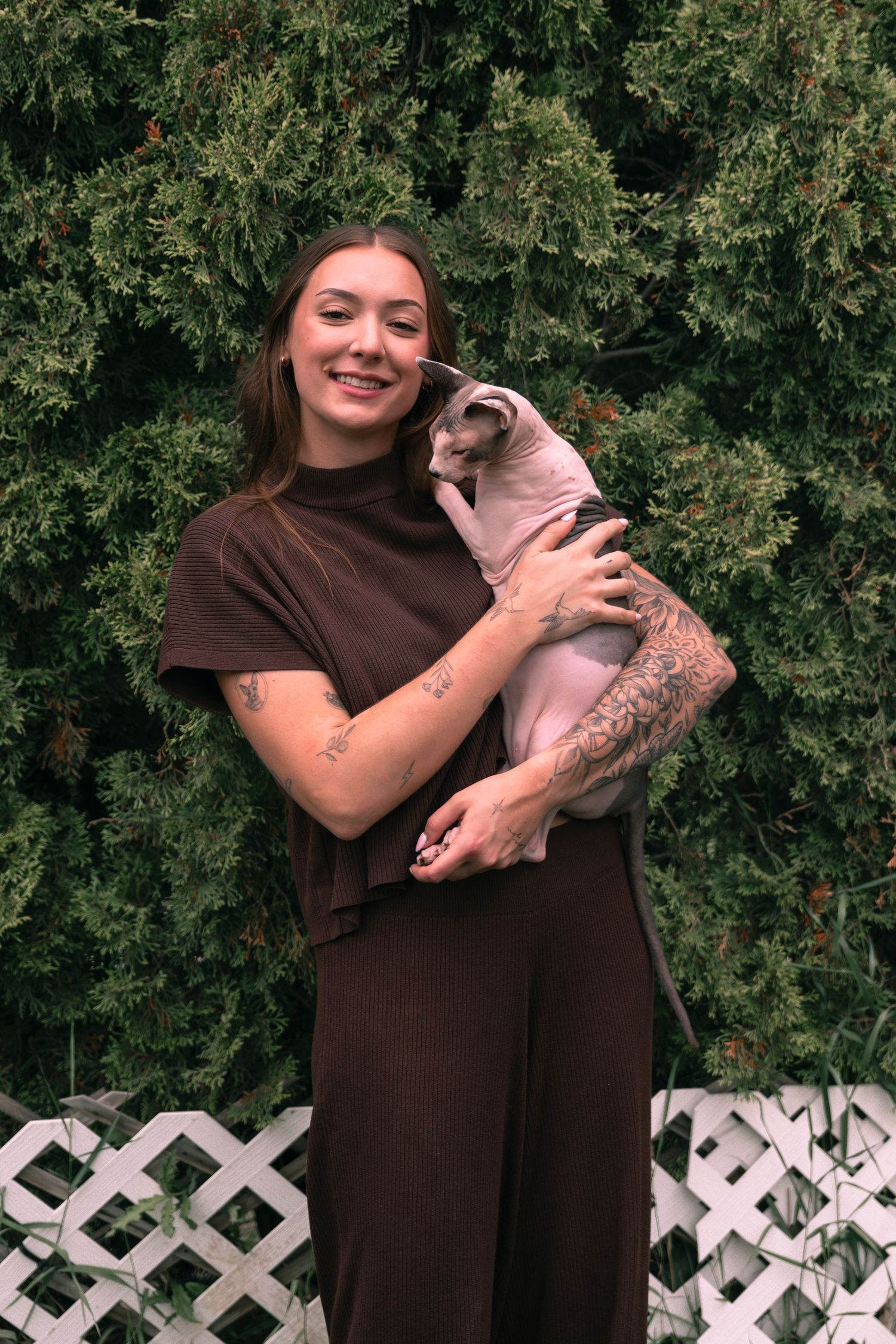 Smiling young woman with tattoos holding a hairless cat outdoors near green shrubs wearing a brown outfit.