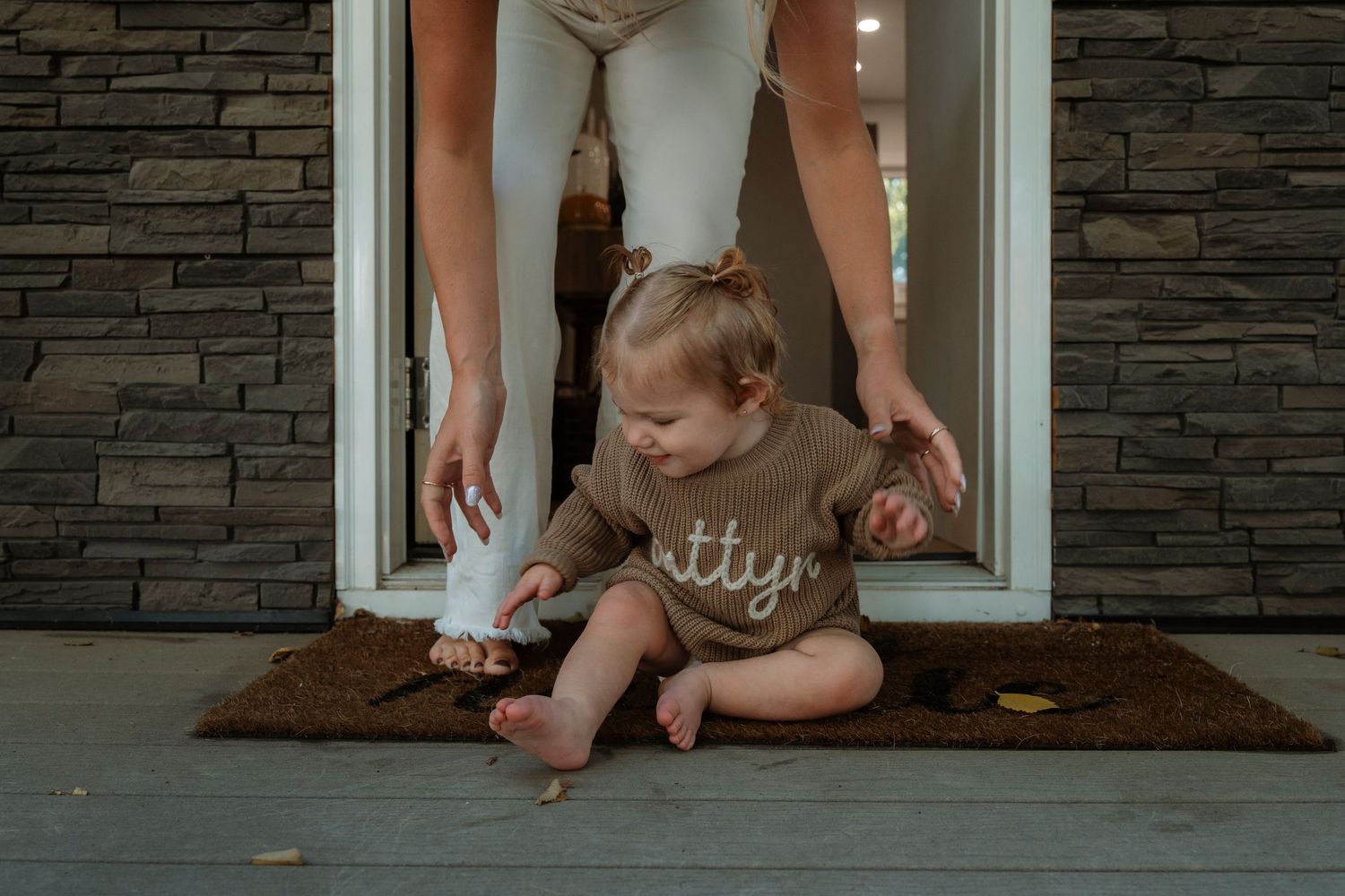 Baby girl in brown sweater sits on doormat holding bottle while adult stands behind in doorway.