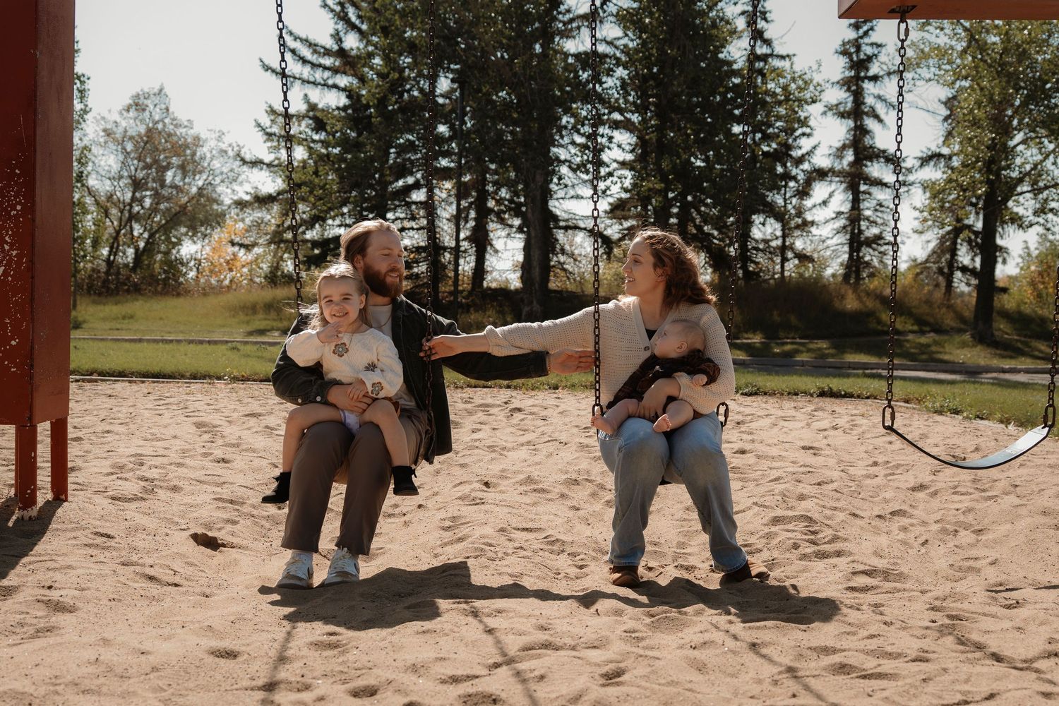 Family with two babies playing on a sandy playground, with tall trees in the background on a sunny day.