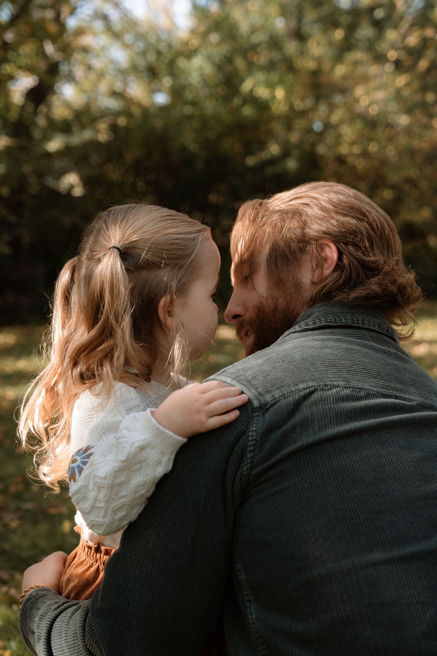 A bearded redhead man tenderly holds a young blonde girl outdoors in a lush green garden setting.