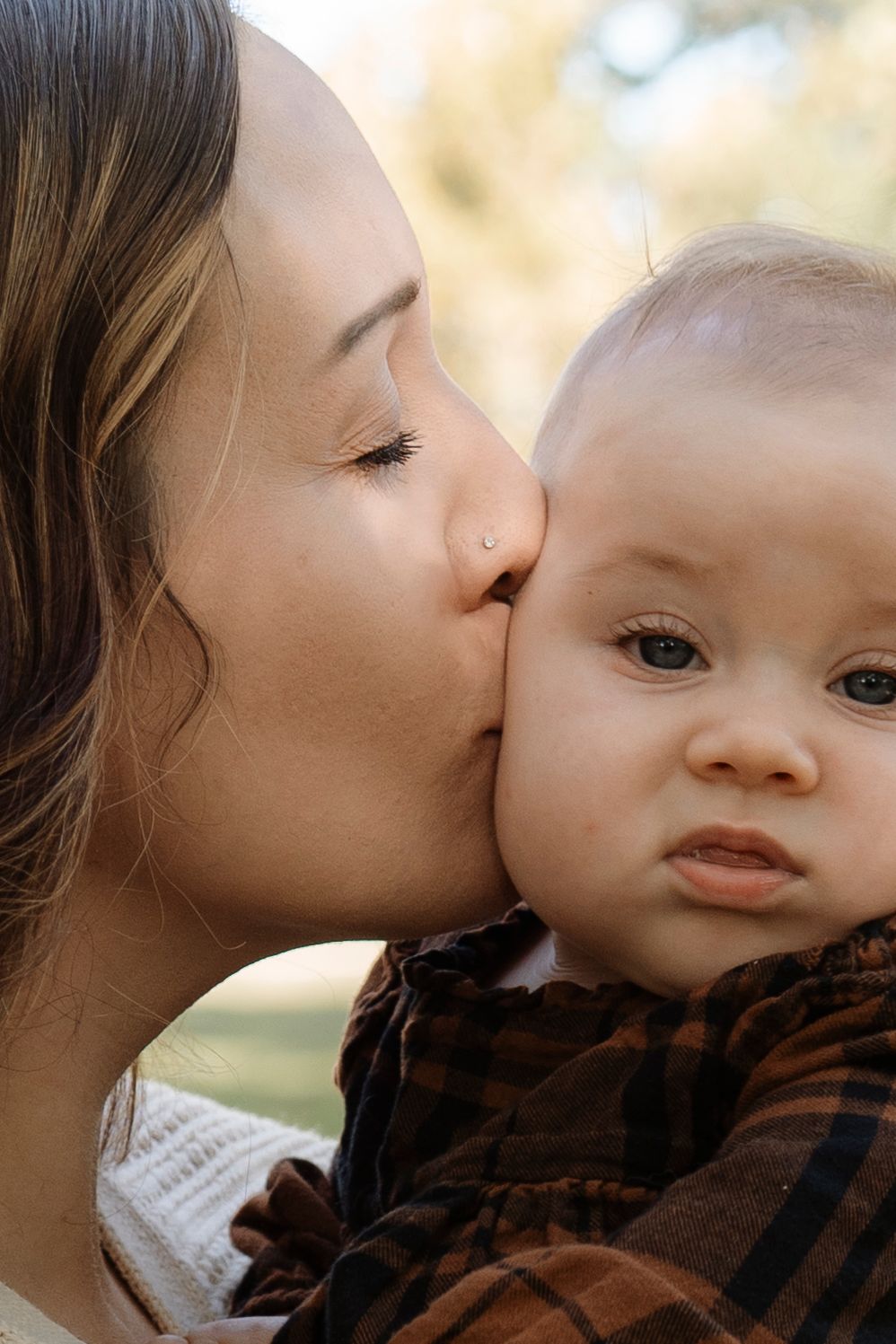A loving mother gently kisses her baby's cheek outdoors, as the infant looks curiously at the camera.