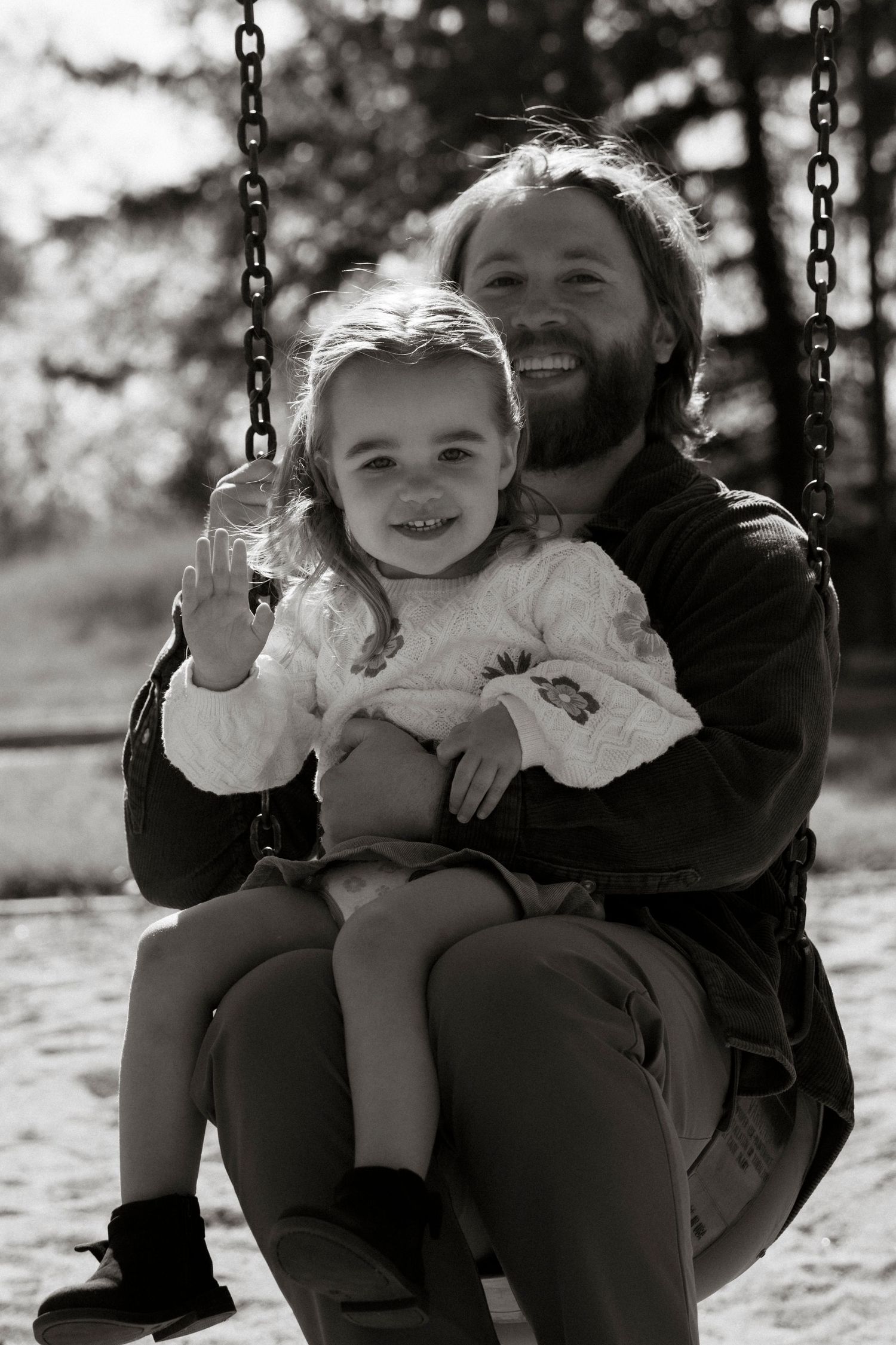 Black and white photo of a bearded father and smiling young daughter sitting together on a playground swing.