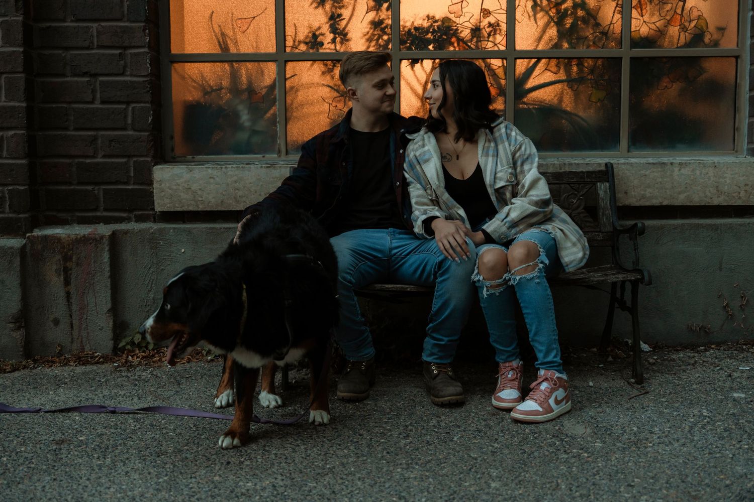 A couple sits on a bench at dusk with their Rottweiler dog beside them, sharing a romantic moment outdoors.