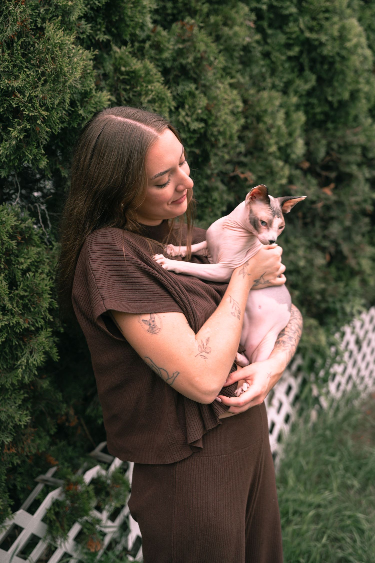 Smiling woman in brown outfit holding a hairless Sphynx cat outdoors near green hedges.