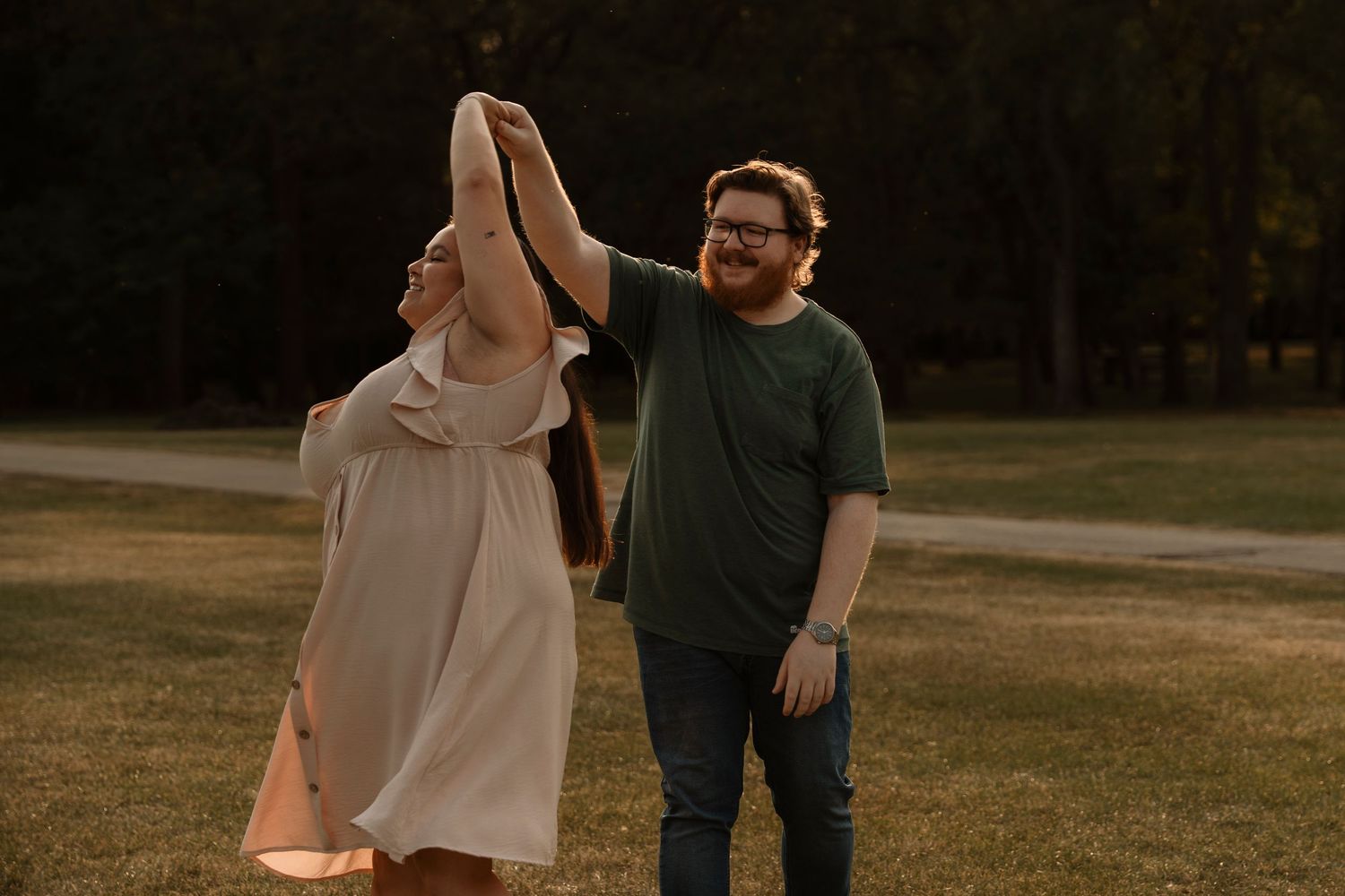 A couple dances outdoors at golden hour, man in green shirt spinning woman in white dress on green grass.