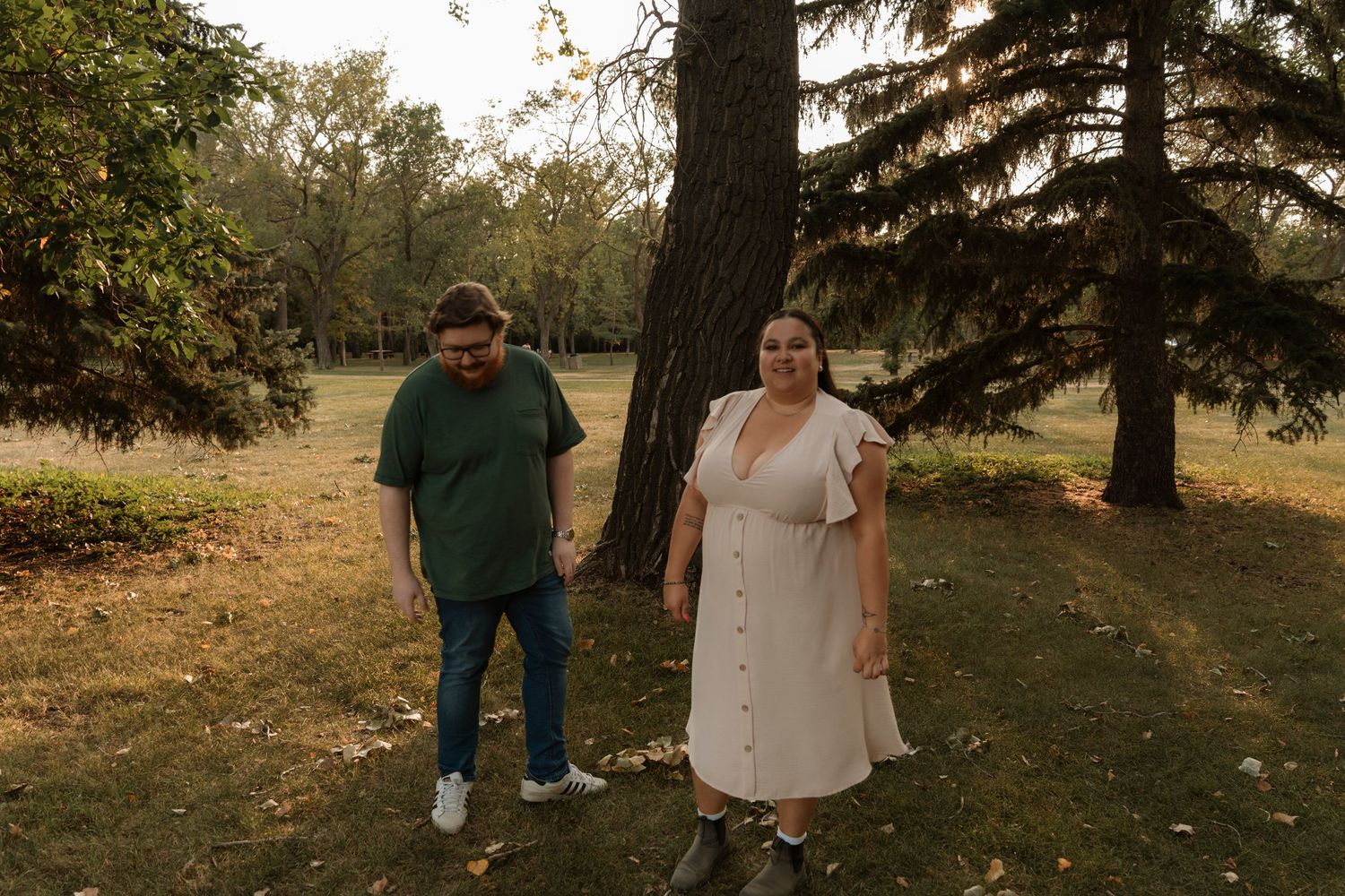 A couple stands apart in a park near pine trees, woman in white dress, man in green shirt and jeans, golden hour light.
