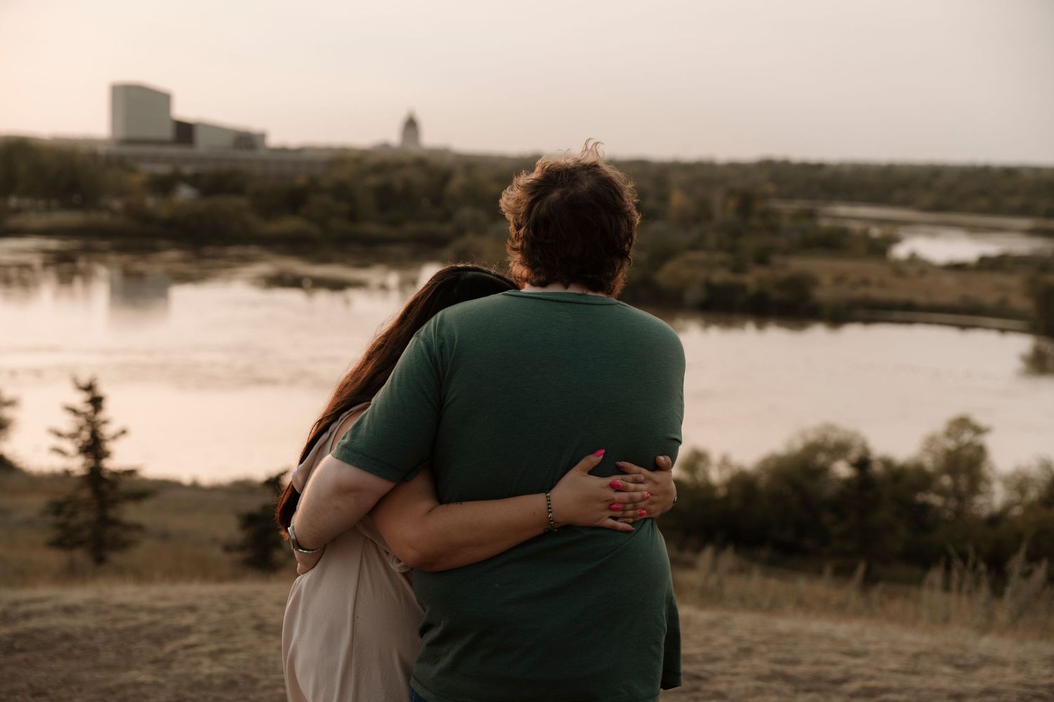 Couple embracing from behind, overlooking a serene river landscape at dusk with trees and a building in the distance.