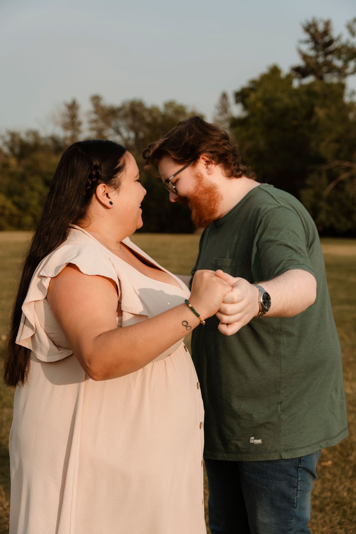 Couple holding hands and smiling at each other outdoors in a sunny park, woman in white dress, man in green shirt.