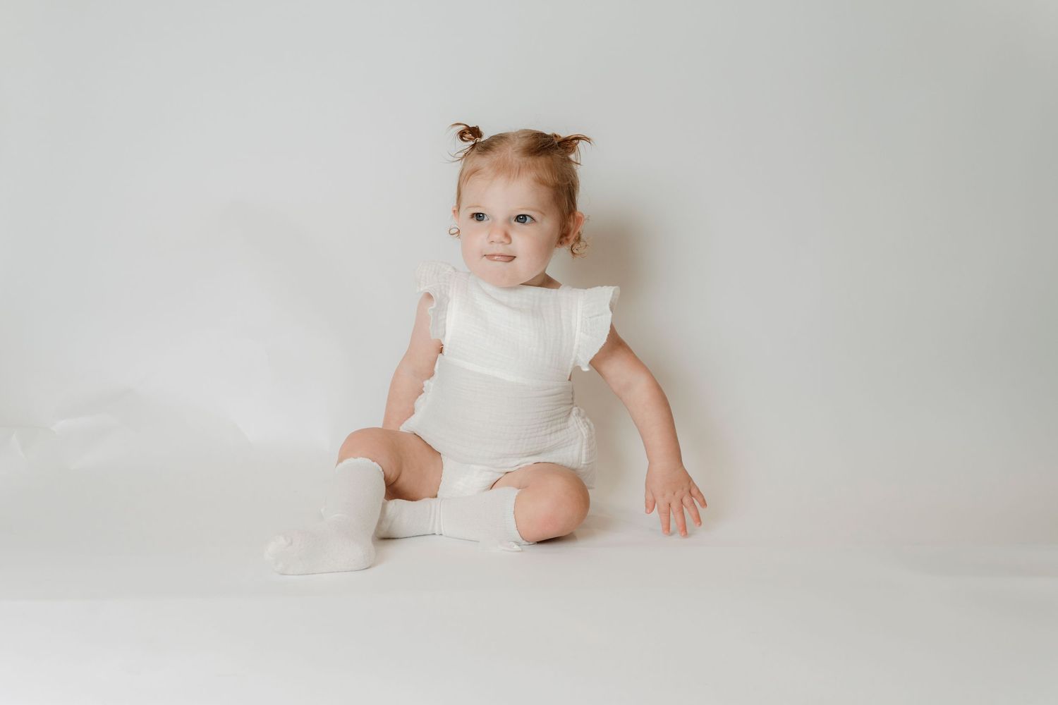 Smiling baby girl in white ruffle romper and socks sitting on white background.