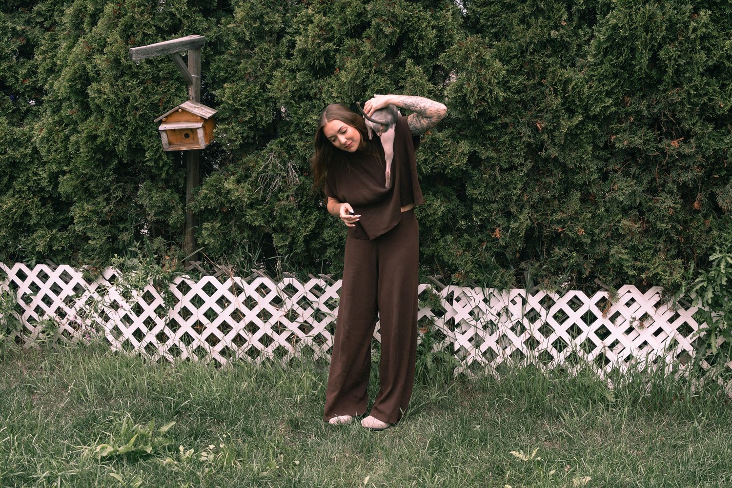 Woman in brown jumpsuit standing on grass near white lattice fence and birdhouse with green hedges backdrop.