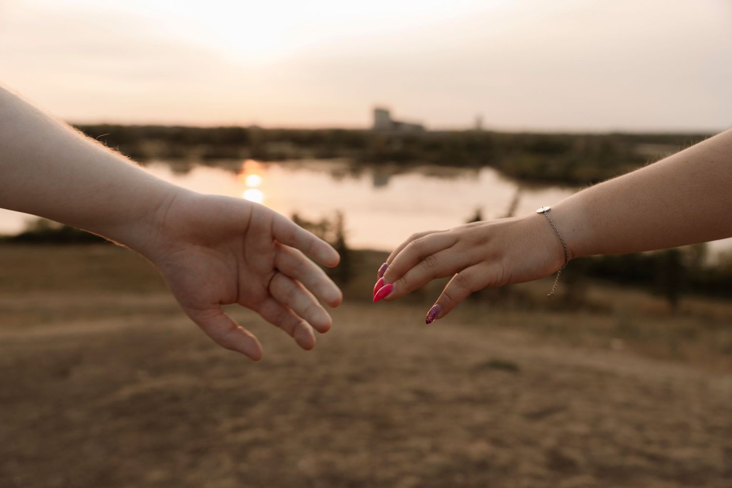 Two hands almost touching at sunset near a waterfront, one with pink nails and a bracelet, in a romantic reaching gesture.